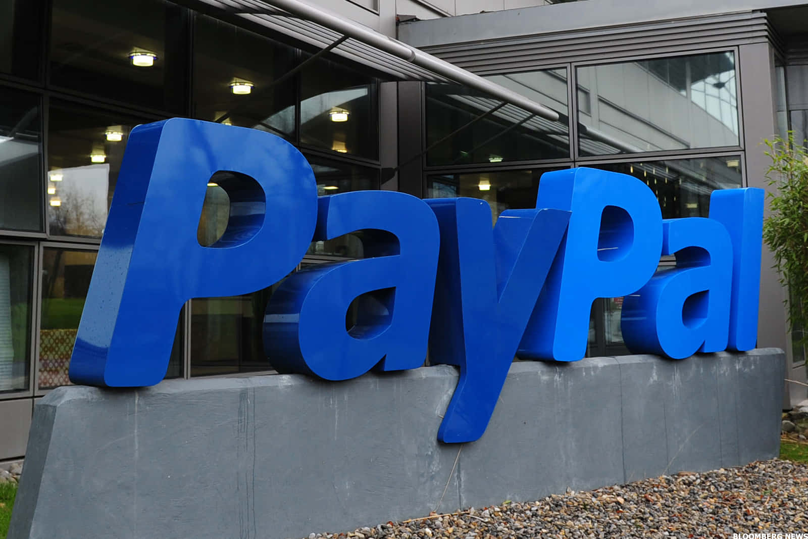 成為全球資金轉移者——PayPal 壁紙 — 大型藍色PayPal標誌字母放置在現代玻璃建築外的灰色底座上，地面鋪有礫石。