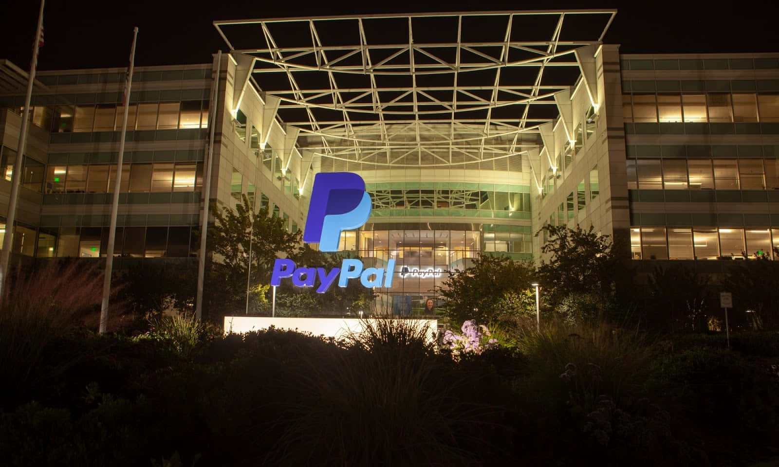 透過PayPal安全收款 壁紙 — 現代玻璃建築的夜景，帶有發光的PayPal標誌和周圍的樹葉。