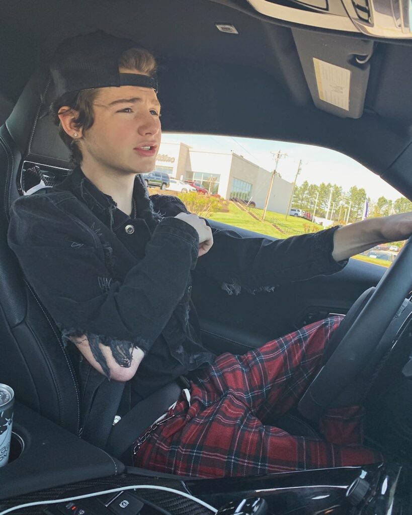 Payton Moormeier 駕車 壁紙 — 一名年輕男子手臂上有紋身，戴著黑色鴨舌帽和紅色格子褲，隨意地坐在一輛時尚黑色汽車的駕駛座上。