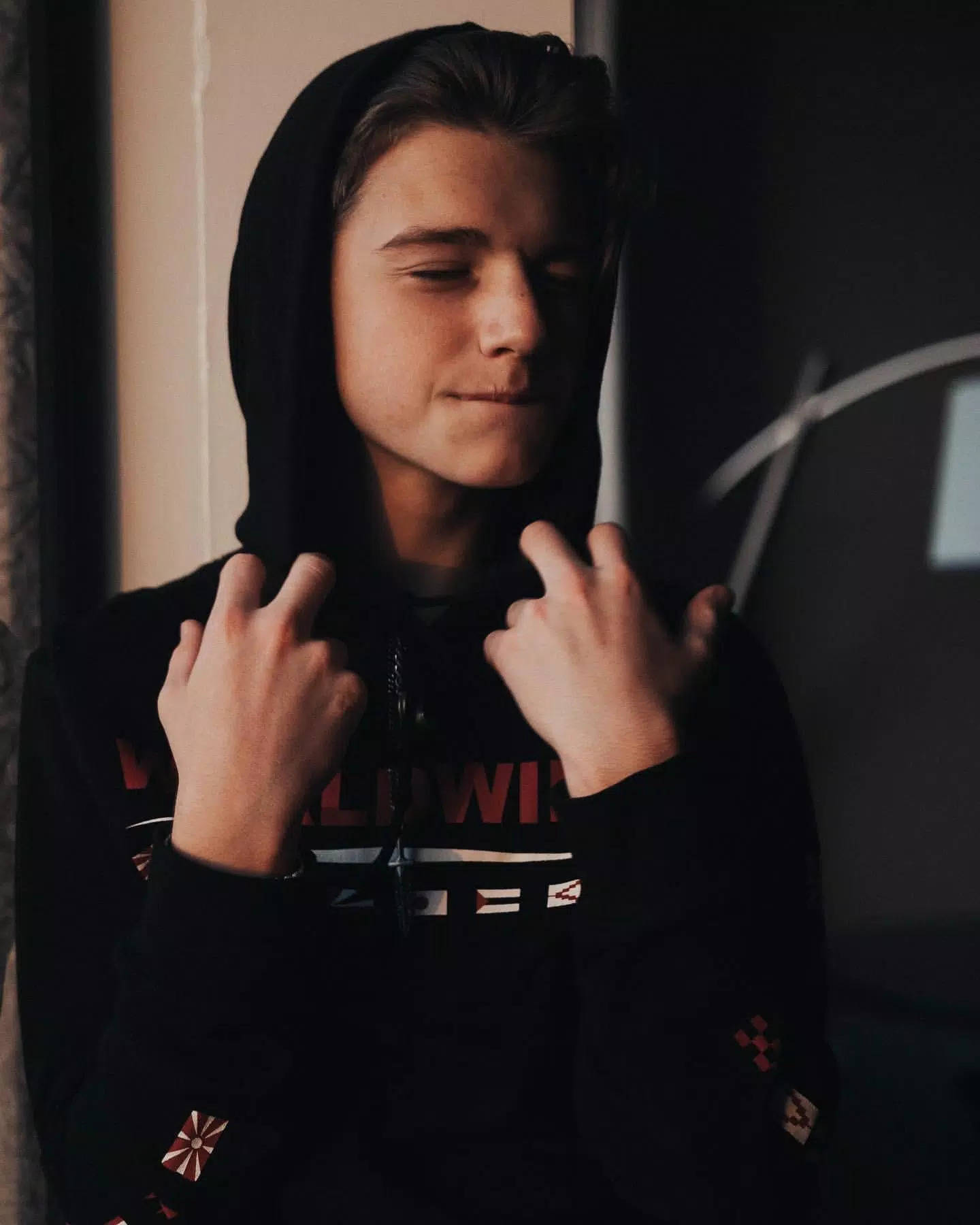 Payton Moormeier 穿黑色連帽衫 壁紙 — 一個年輕男子穿著黑色連帽衫，閉著眼睛，雙手調整帽子，在昏暗的房間裏。