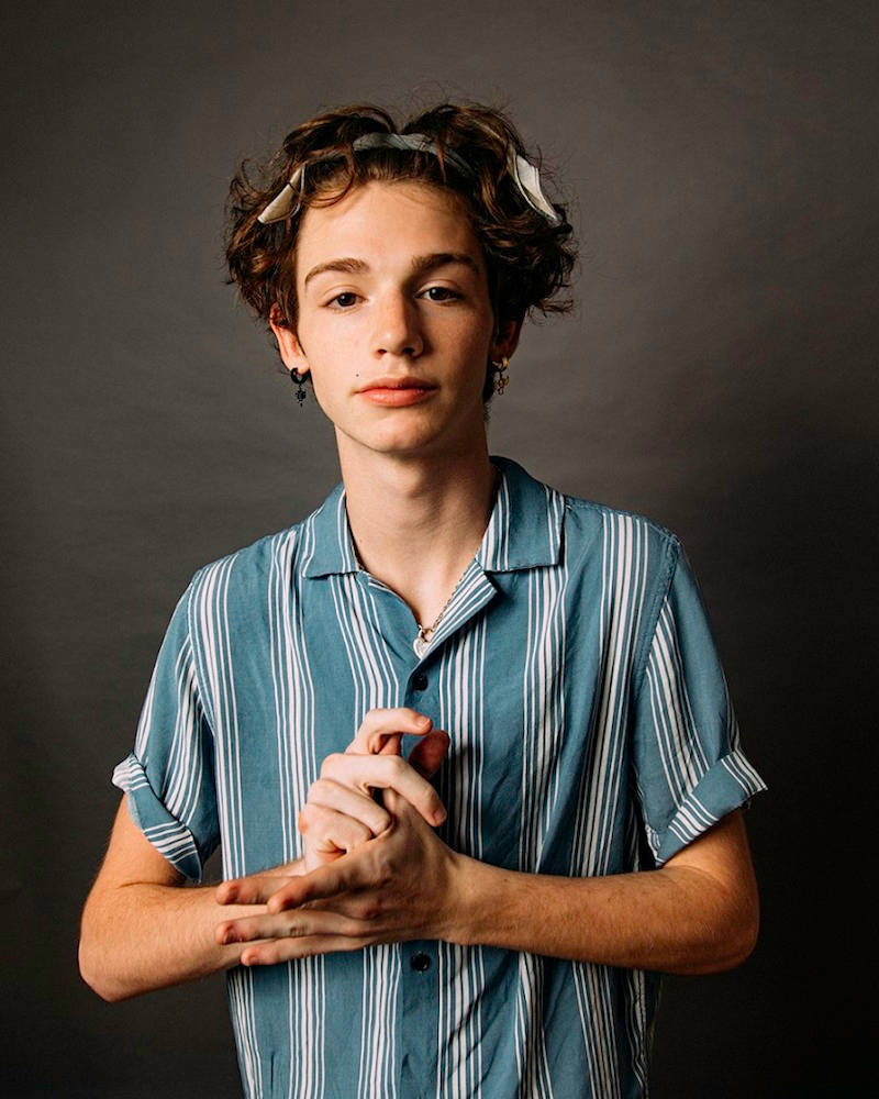 Payton Moormeier 穿藍色襯衫 壁紙 — 一個年輕人，頭髮捲曲，眼鏡架在頭上，穿著藍色條紋襯衫，背景是深灰色攝影棚。