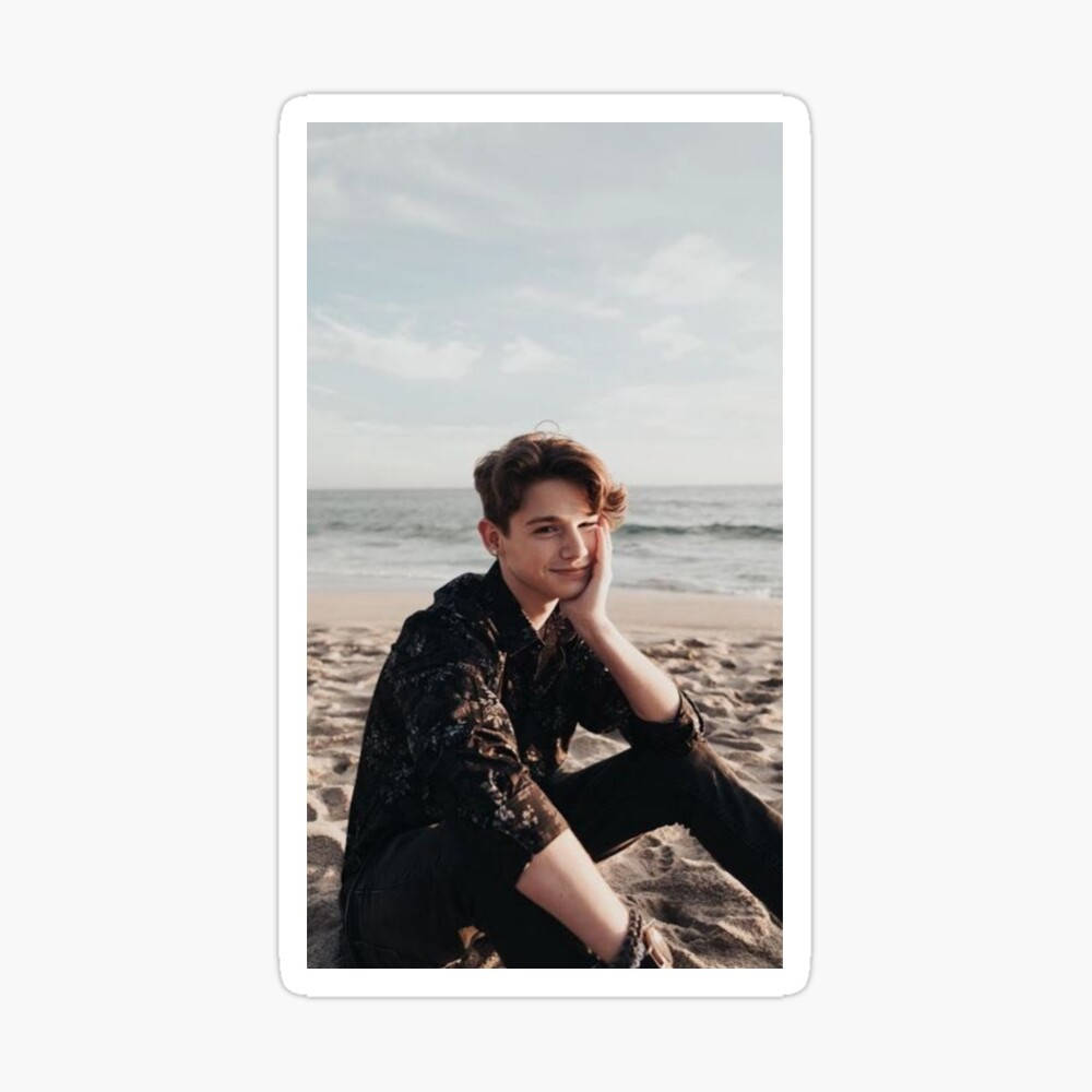 Payton Moormeier 海灘肖像 壁紙 — 一位年輕男子留著波浪形棕色頭髮，坐在沙灘上，手托著臉頰溫柔微笑，身後的海浪在柔和多雲的天空下翻滾。