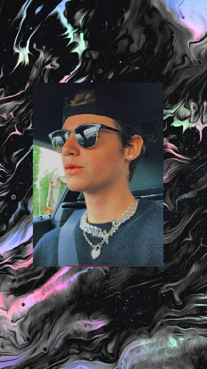 Payton Moormeier 在車內 壁紙 — 一個人戴著太陽眼鏡和冷帽坐在車內，背景是帶有粉紅、紫色和藍綠色調的旋轉宇宙星雲。