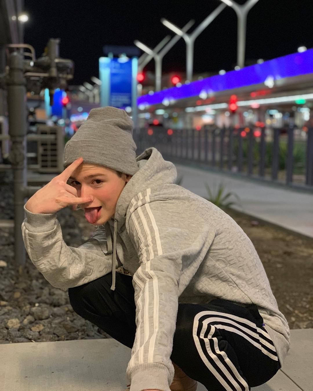 Payton Moormeier 穿著舒適服裝 壁紙 — 一個穿著灰色連帽衫和冷帽的年輕人，在夜晚頑皮地蹲下，伸出舌頭並做出手勢，背景是燈火通明的城市。