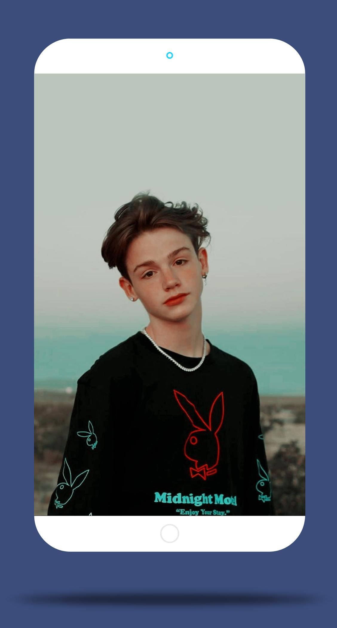 Payton Moormeier 手機風格編輯 壁紙 — 一位年輕人在智能手機屏幕上，穿著黑色圖案T恤，背景是朦朧的海岸，頭髮造型時尚，戴著耳環。