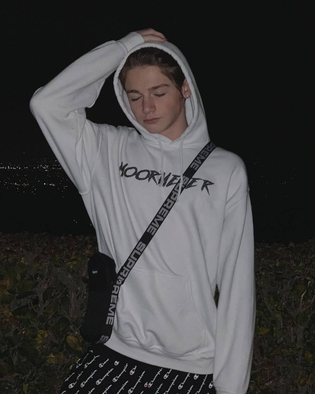 Payton Moormeier 穿著自家商品 壁紙 — 一個穿著印有 'MOORWATER' 字樣白色連帽衫的年輕人，在夜晚站在黑暗的天空下，下方是遠處的城市燈光。