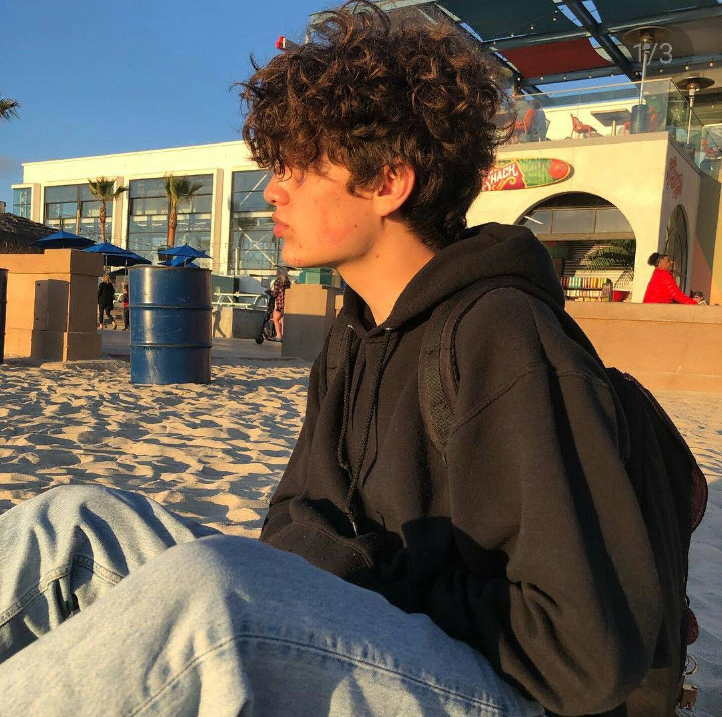 Payton Moormeier 金色時光 壁紙 — 一個年輕人留著捲髮，穿著黑色連帽衫和牛仔褲坐在沙灘上，金色陽光灑在他們的側臉上。