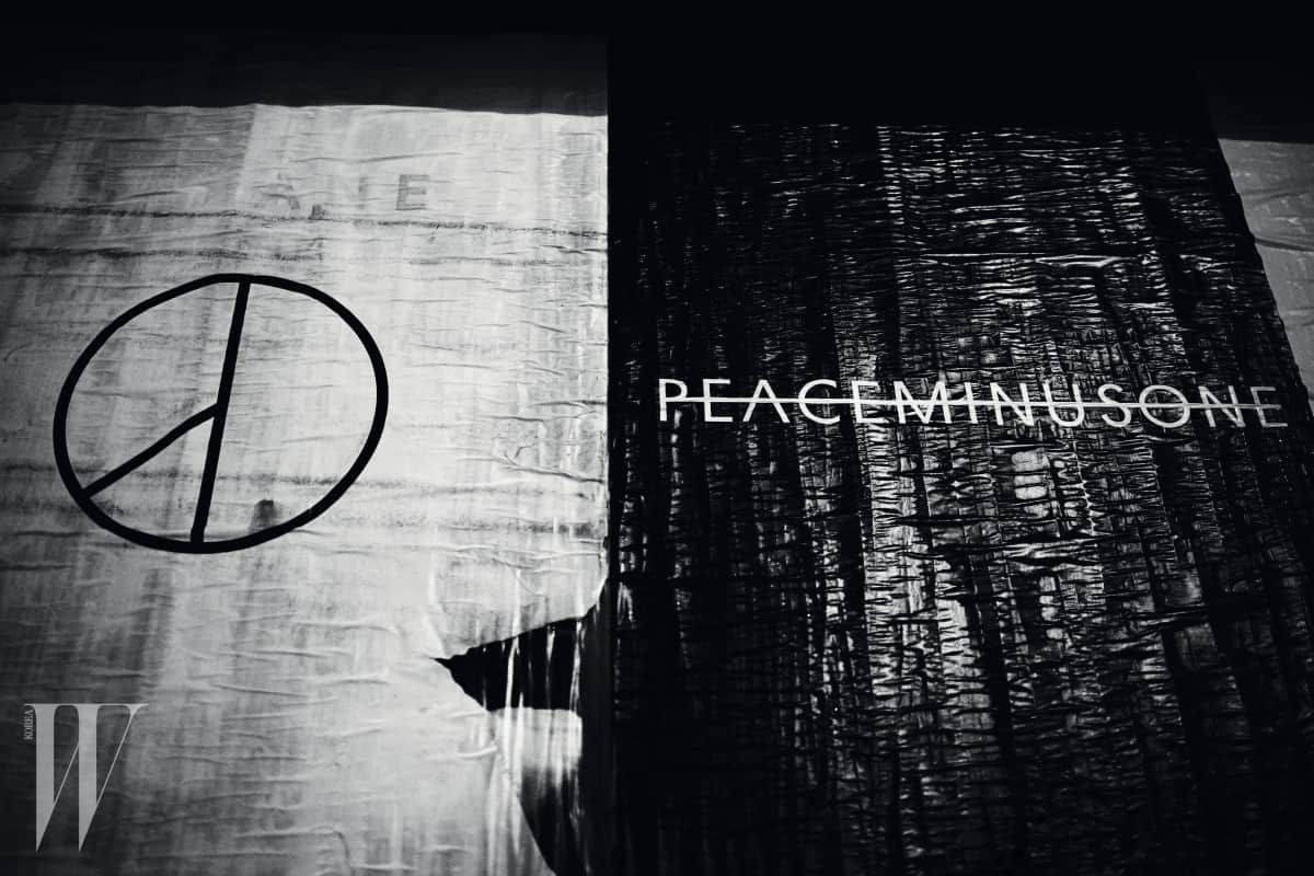PEACEMINUSONE 奢華品牌 壁紙 — 一張清晰的黑白照片，在戲劇性燈光下，紋理布料上顯示和平符號和「PEACEMINUSONE」文字。