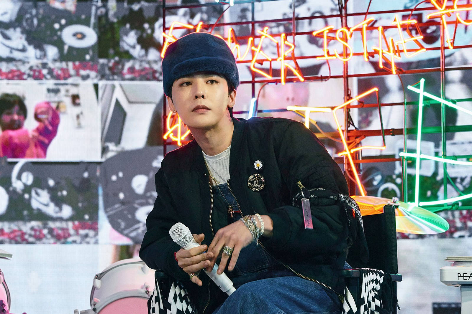 Peaceminusone 與 Kwon Ji Yong 壁紙 — 一個戴藍色冷帽、穿黑色外套的年輕男子，在充滿霓虹燈光、塗鴉和照片的背景下拿著麥克風。