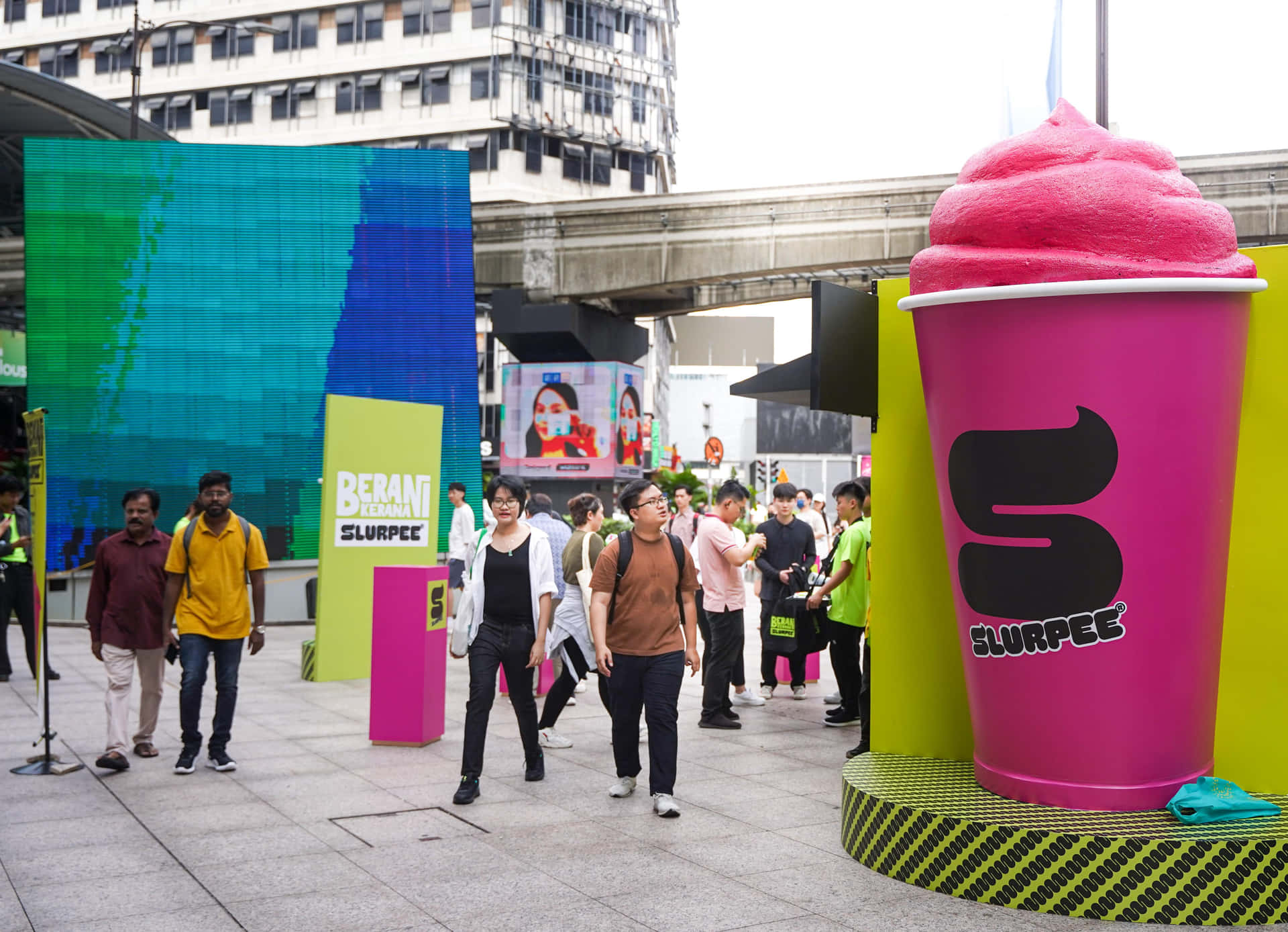  壁紙 — 行人漫步經過一個巨大的粉紅色 Slurpee 杯雕塑和色彩繽紛的品牌裝置，位於繁華的城市廣場。