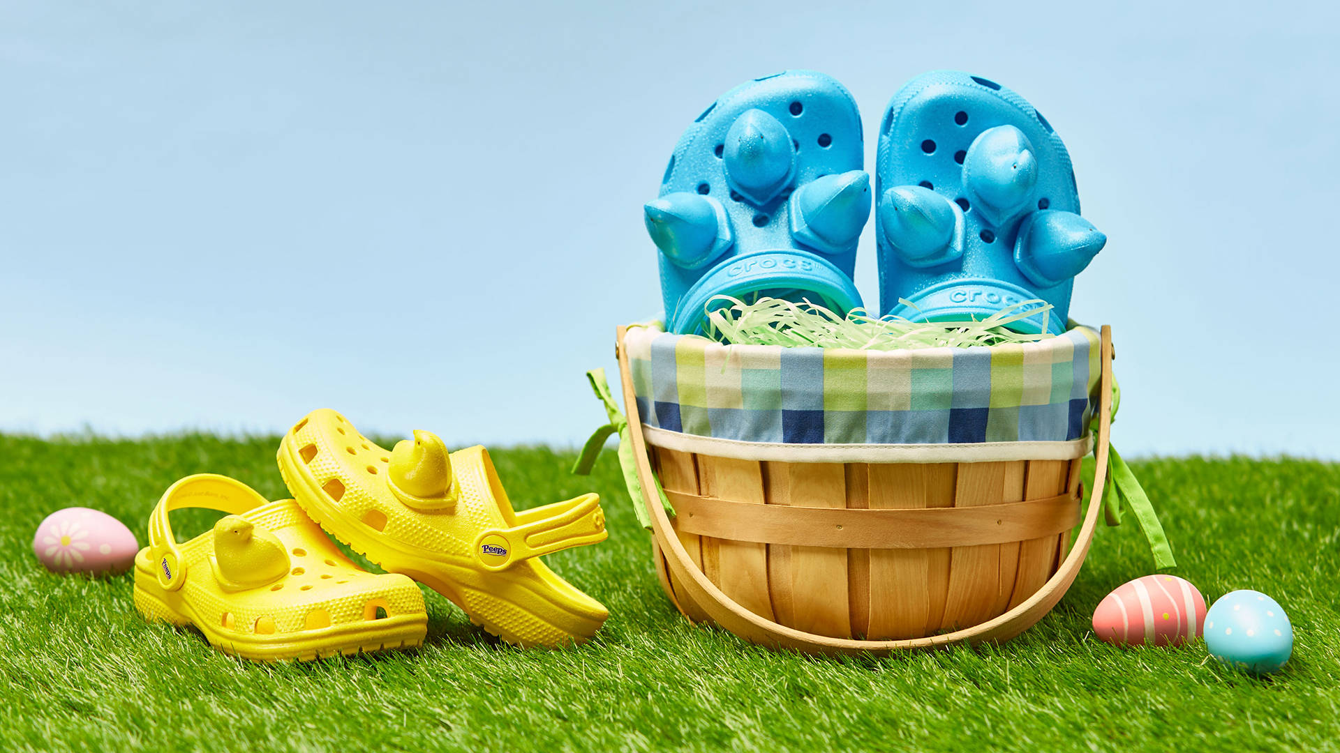 Peeps x Crocs經典款 壁紙 — 藍色和黃色的Crocs涼鞋放在一個編織籃子裡，裡面有復活節彩蛋，放在充滿活力的綠色草地上，天空晴朗。