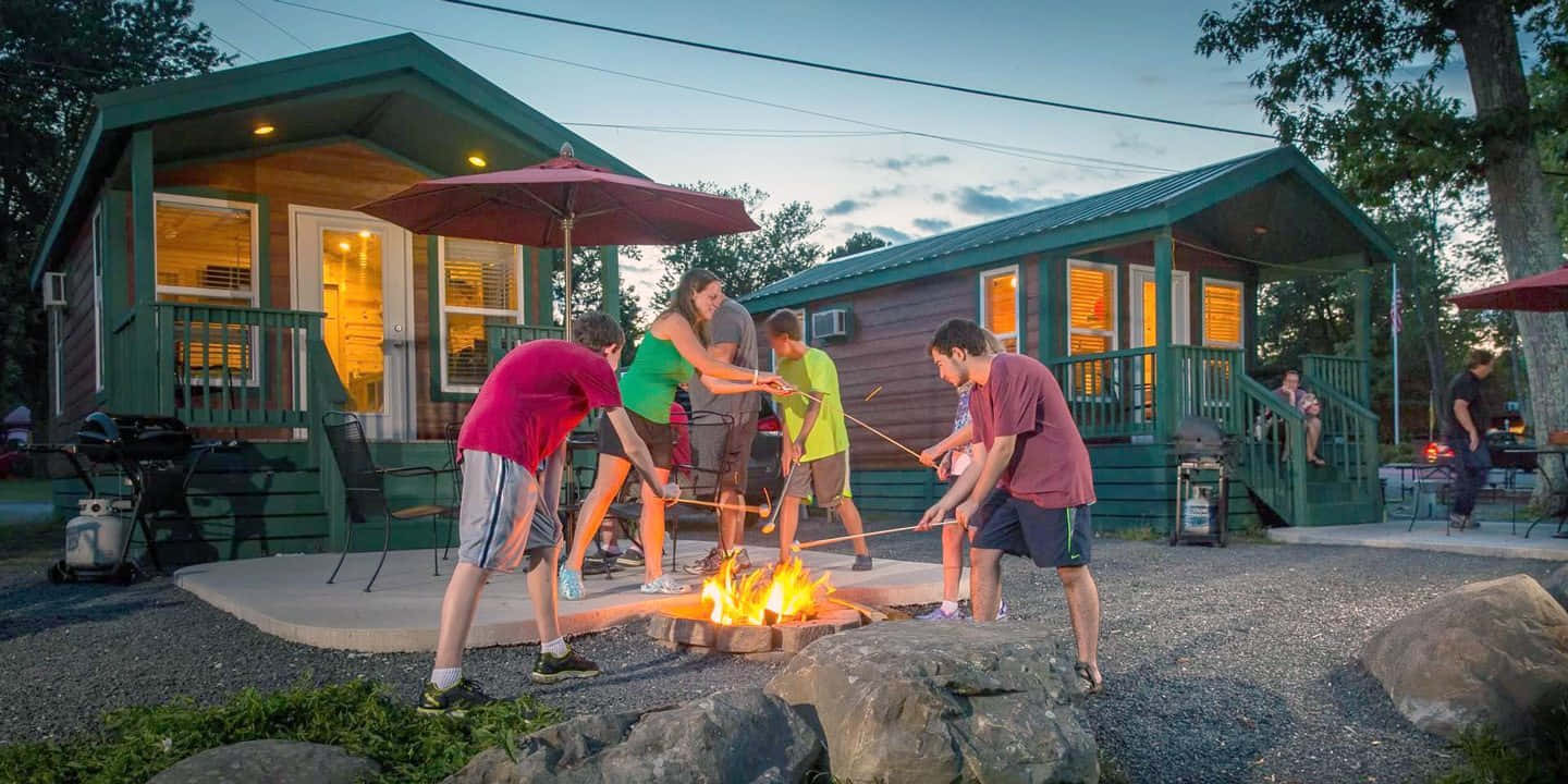 Fall Camping 桌布 壁紙 — 朋友們在黃昏時分，在綠色小屋前，圍著營火烤棉花糖，屋內透出溫暖的燈光。
