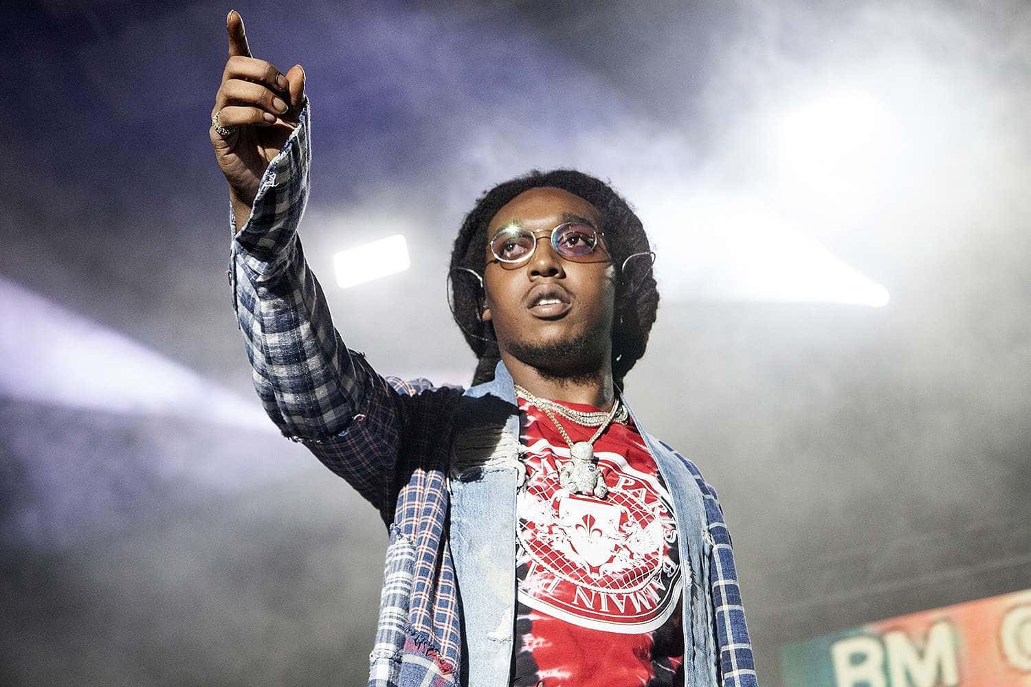  壁紙 — 一位戴眼鏡和辮子頭的表演者在煙霧彌漫的舞台上，在明亮的聚光燈照射下向上指。