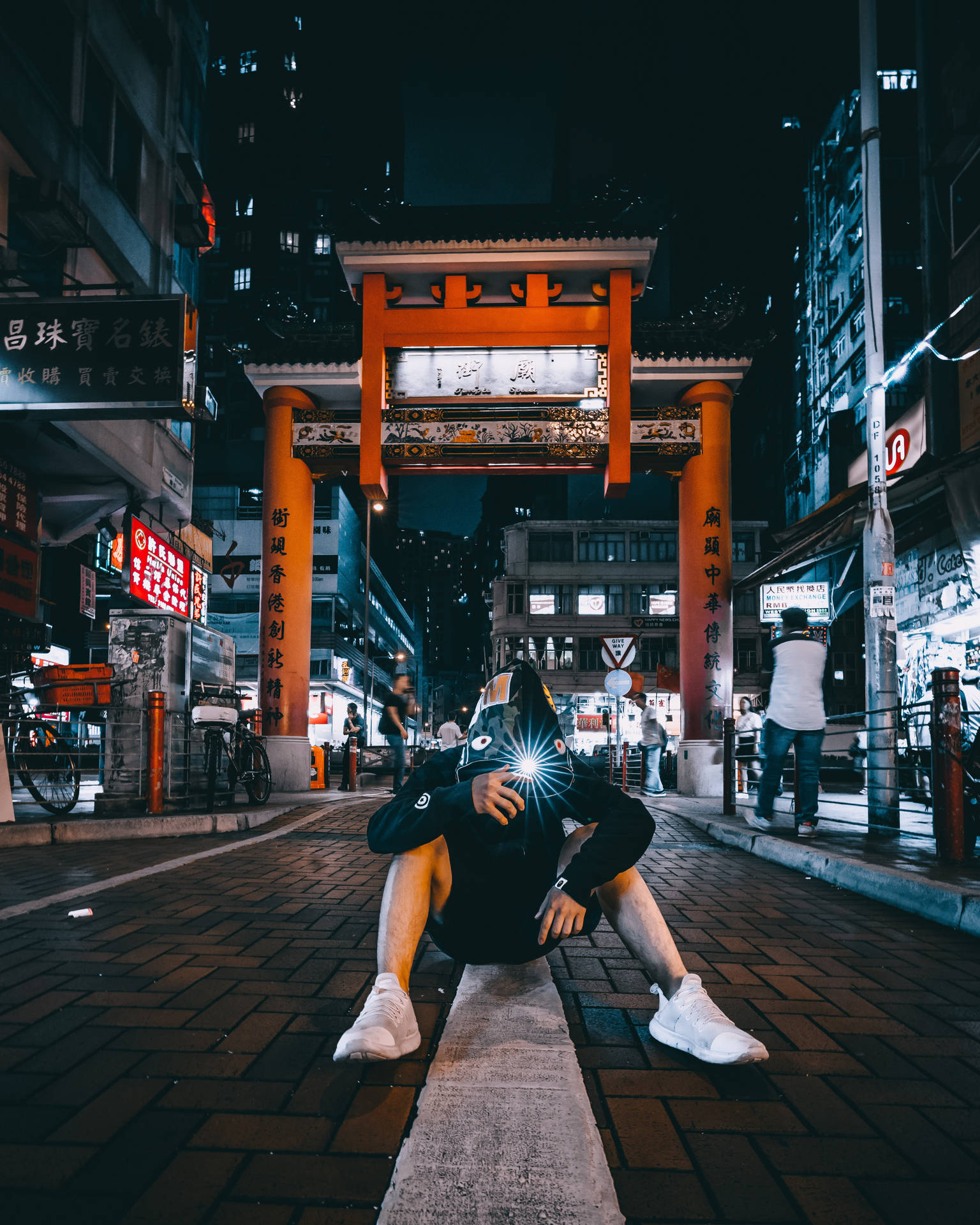 於都市中推動變革 壁紙 — 一個人夜晚蹲喺城市街道上，喺傳統橙色門下揸住發光電話，周圍有霓虹燈牌發光。