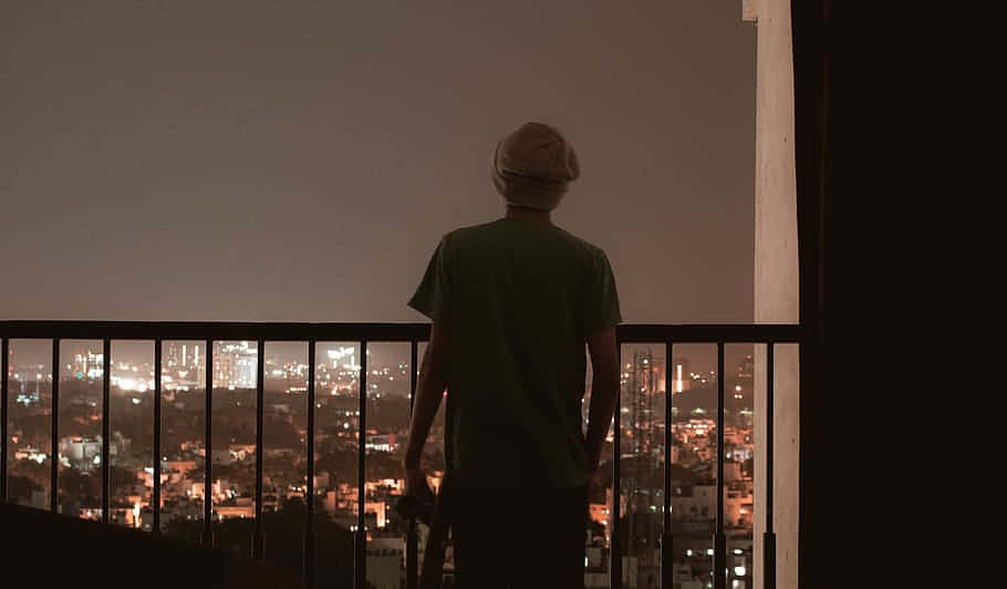 HD Aesthetic 桌布 壁紙 — 一個人戴著冷帽，夜晚獨自站在陽台上，俯瞰著被溫暖燈光照亮的廣闊城市景觀。