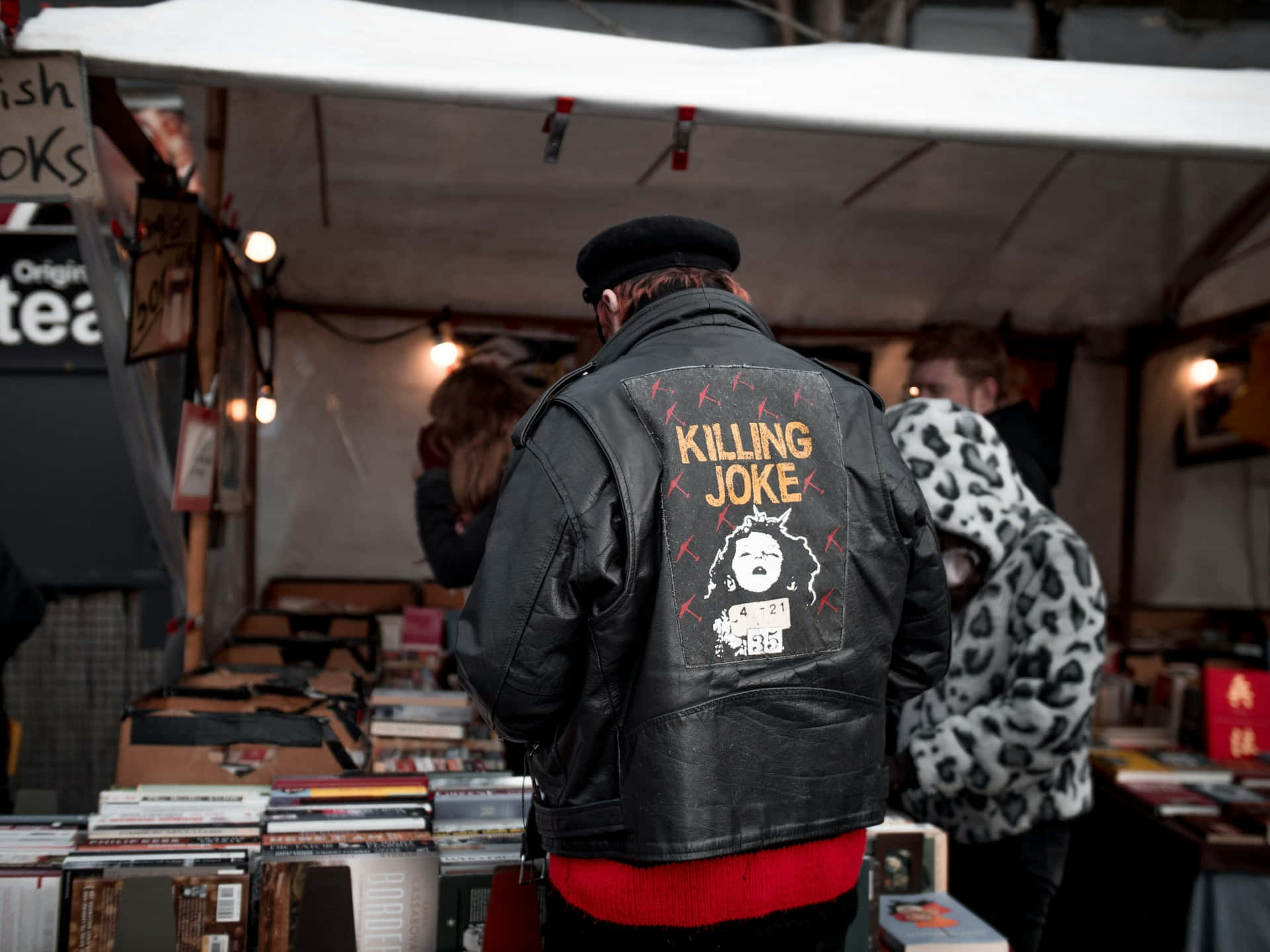  壁紙 — 一個穿著印有「Killing Joke」圖案黑色皮褸的人，站在白色帳篷下一個熙來攘往的書攤旁。