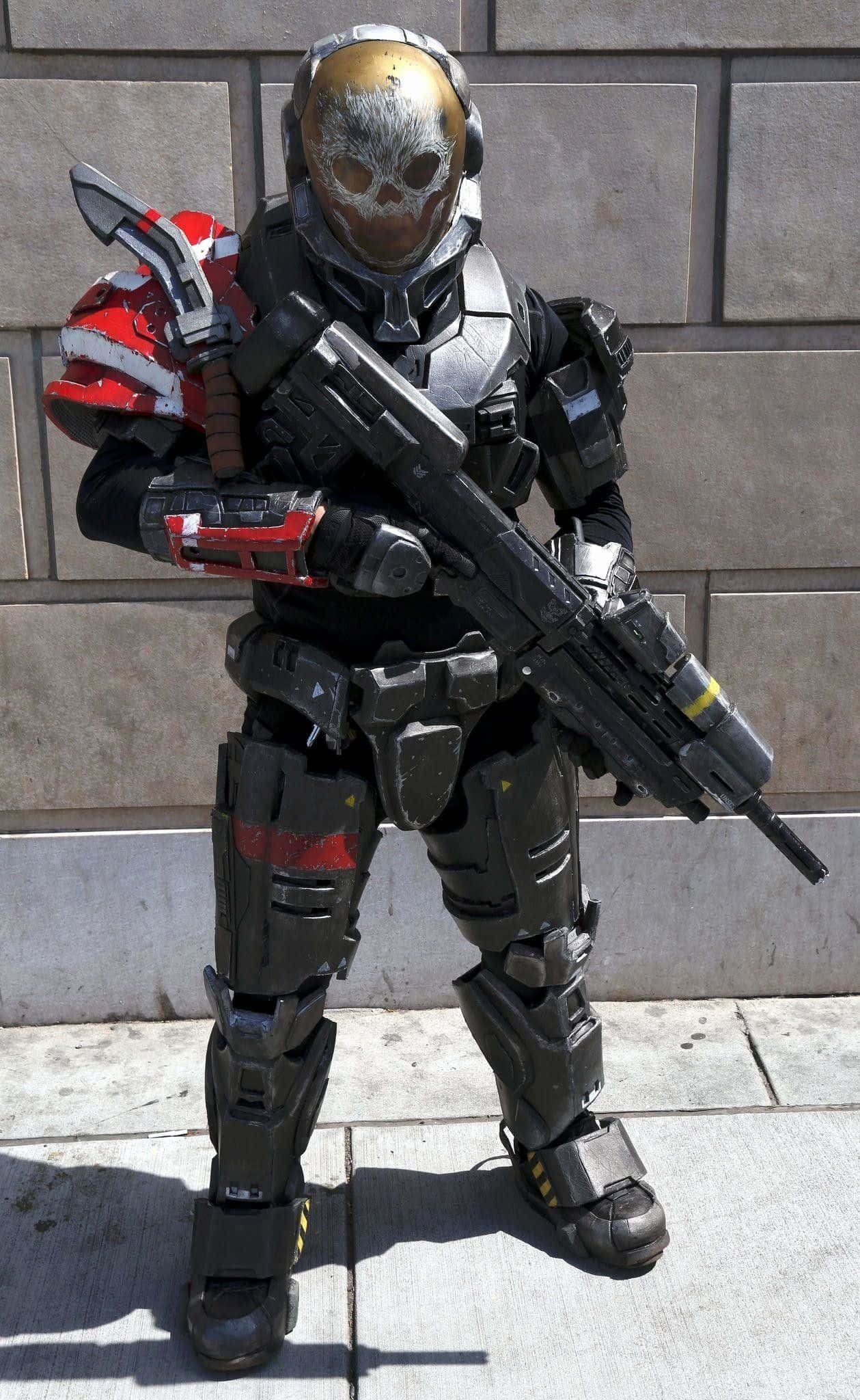 Halo Emile 桌布 壁紙 — 一個穿著精細黑色和紅色盔甲角色扮演、戴著骷髏頭盔、手持步槍的人，站在石牆旁的人行道上。