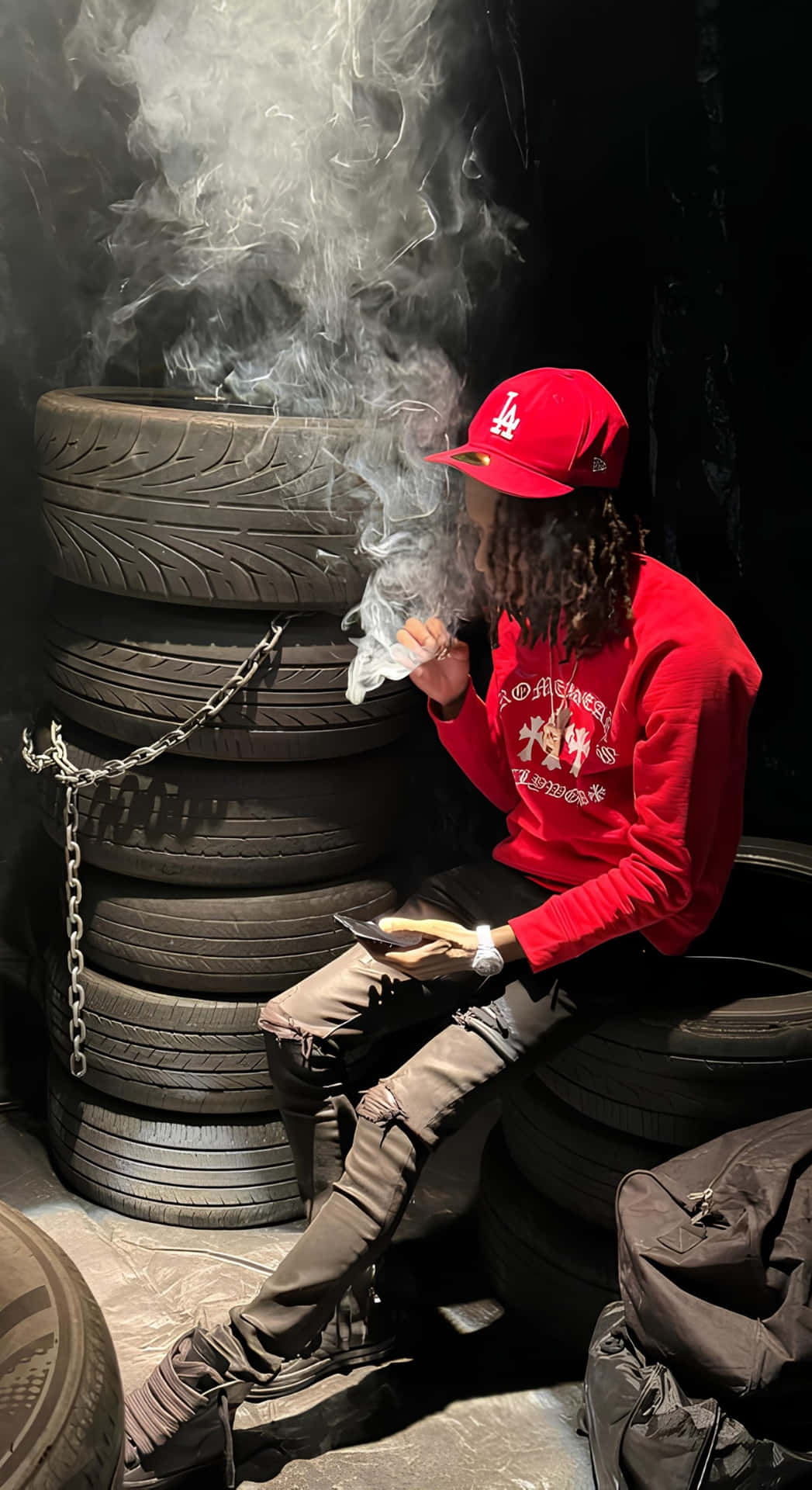  壁紙 — 一個戴住紅色LA帽同連帽衫嘅人坐喺疊起嘅輪胎上面，一邊食煙一邊揸住電話，喺燈光昏暗嘅車房入面。