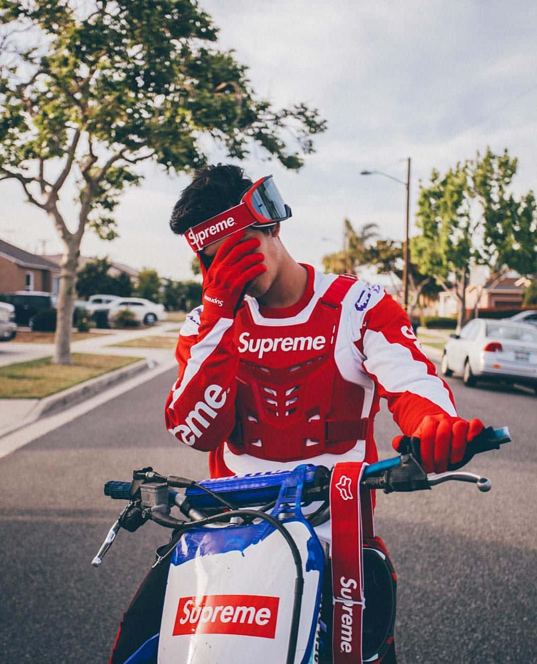 穿著紅色 Supreme 裝備的人，在陽光明媚的郊區街道上，在品牌滑板車上調整護目鏡。