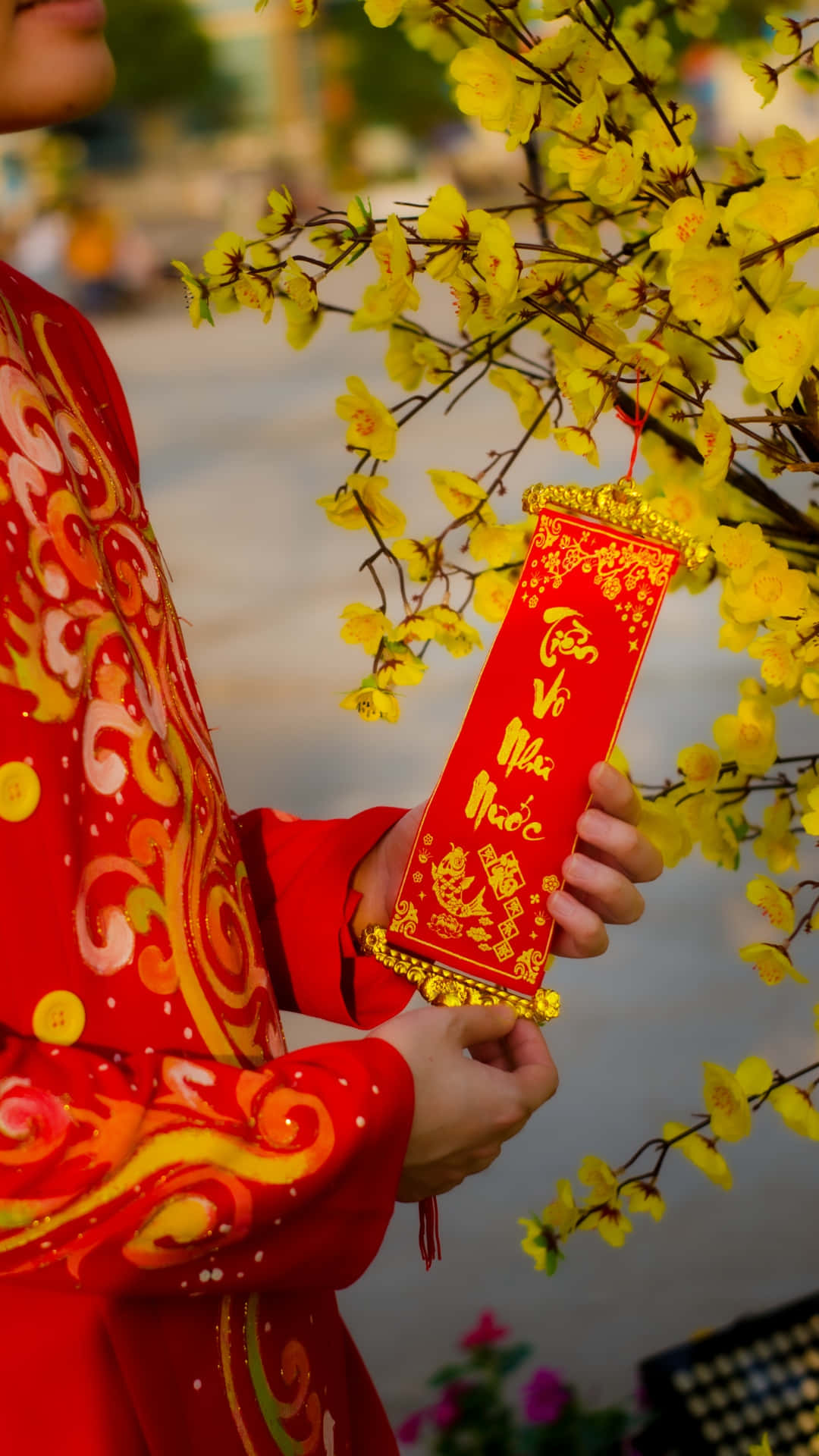  壁紙 — 一位身穿鮮紅色傳統服飾的人，在盛開的黃色花朵下拿著一個金色邊緣的紅色利是封。
