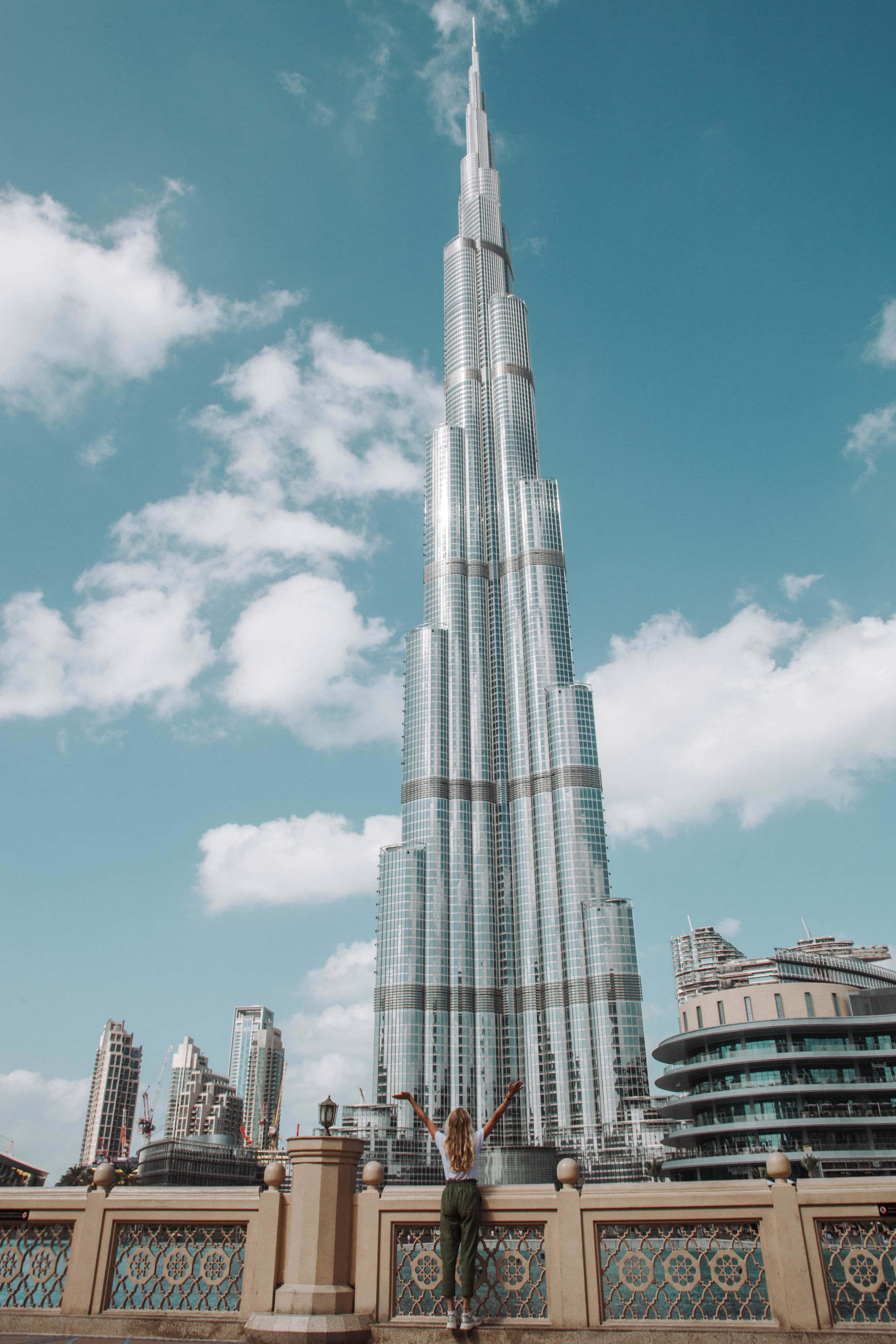 Burj Khalifa 桌布 壁紙 — 一個人舉起雙臂，站在陽台上，俯瞰高聳的哈利法塔，背景是明亮的藍天和蓬鬆的白雲。