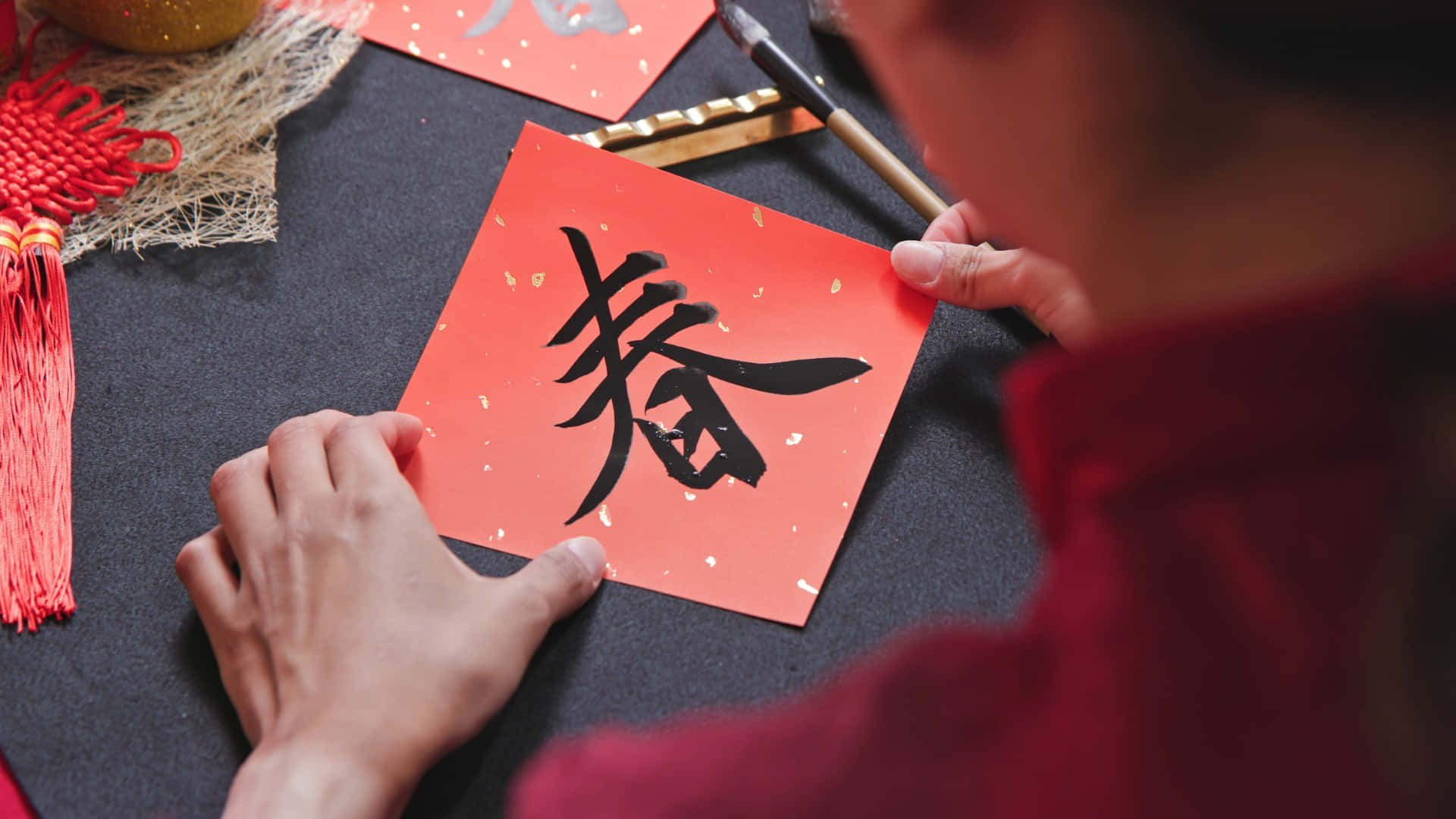  壁紙 — 一個人正在鮮紅色的紙上，用粗黑墨水仔細書寫春天的漢字，周圍環繞著節日裝飾。