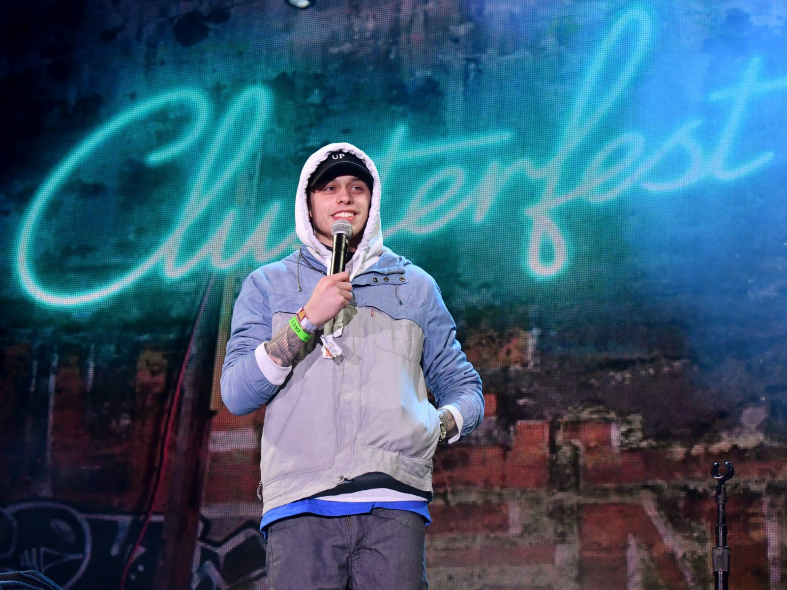 Pete Davidson Clusterfest 單口喜劇表演 壁紙 — 一位微笑的表演者穿著連帽衫，在發光的Clusterfest標誌下拿著麥克風在舞台上。