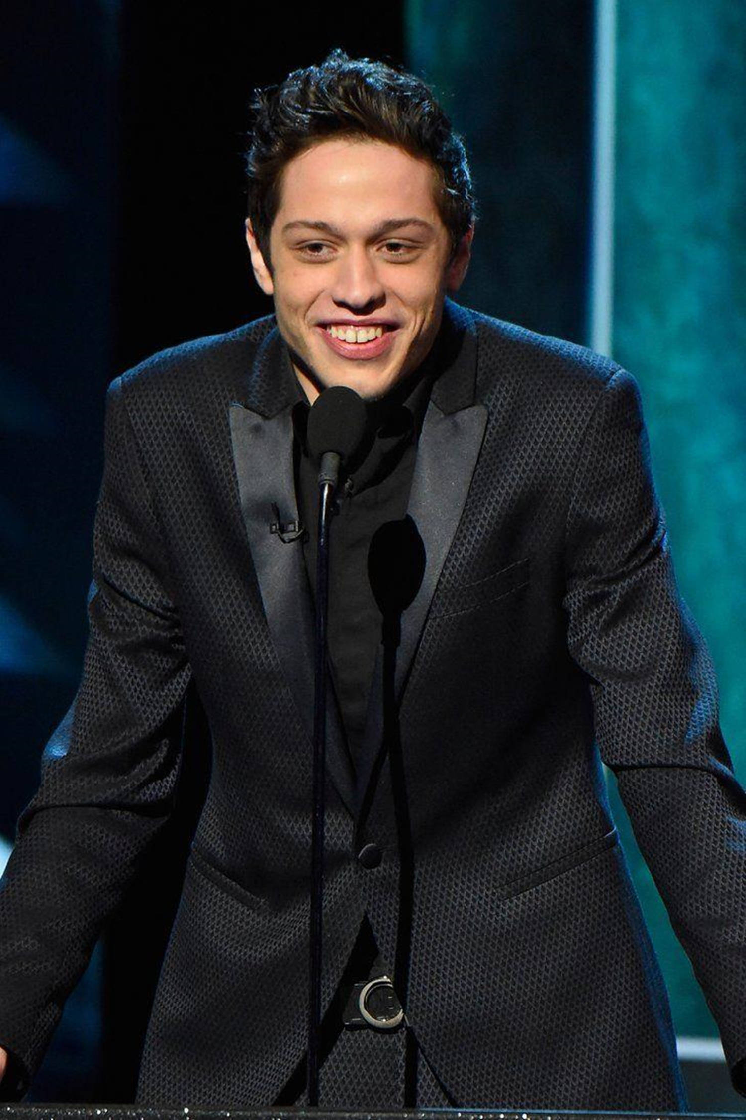 Pete Davidson 評論會 Justin Bieber 壁紙 — 一位面帶微笑、身穿質感黑色西裝的男子，站在燈光昏暗、背景為青綠色的舞台上，面前是講台。