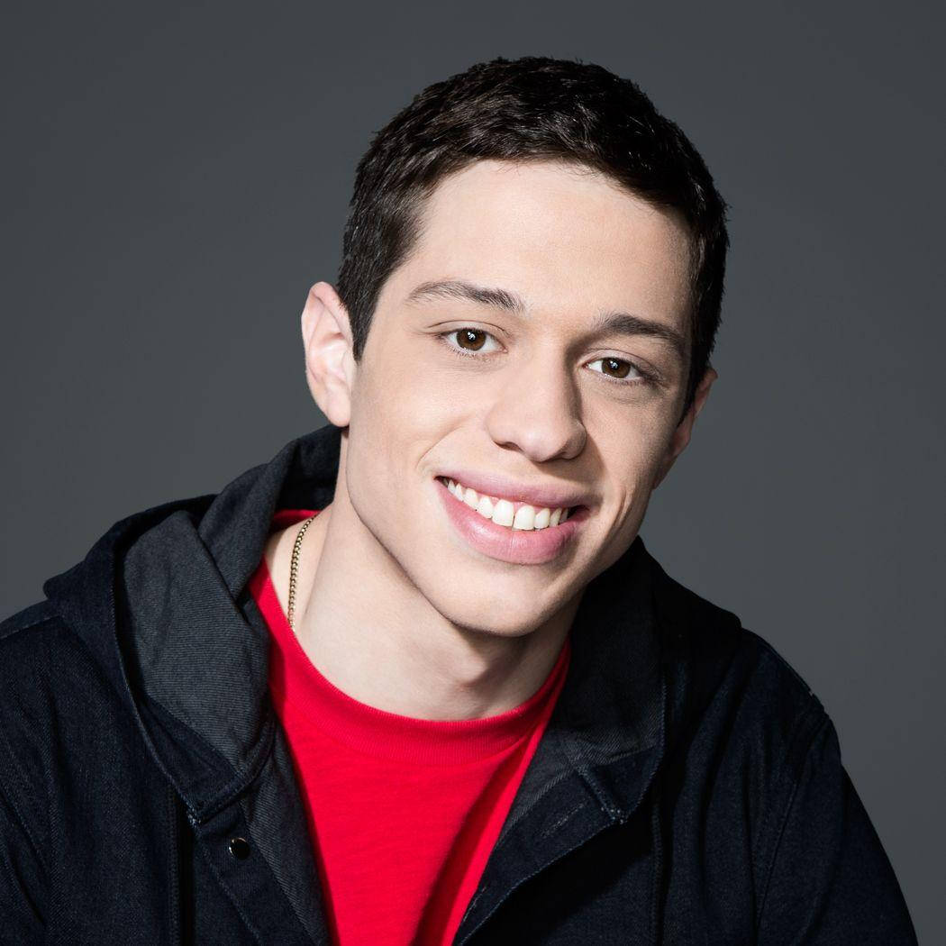 Pete Davidson SNL NBC 廣告拍攝 壁紙 — 一位短黑髮的年輕男子，在灰色背景前穿著黑色連帽衫下的紅色襯衫，溫暖地微笑。