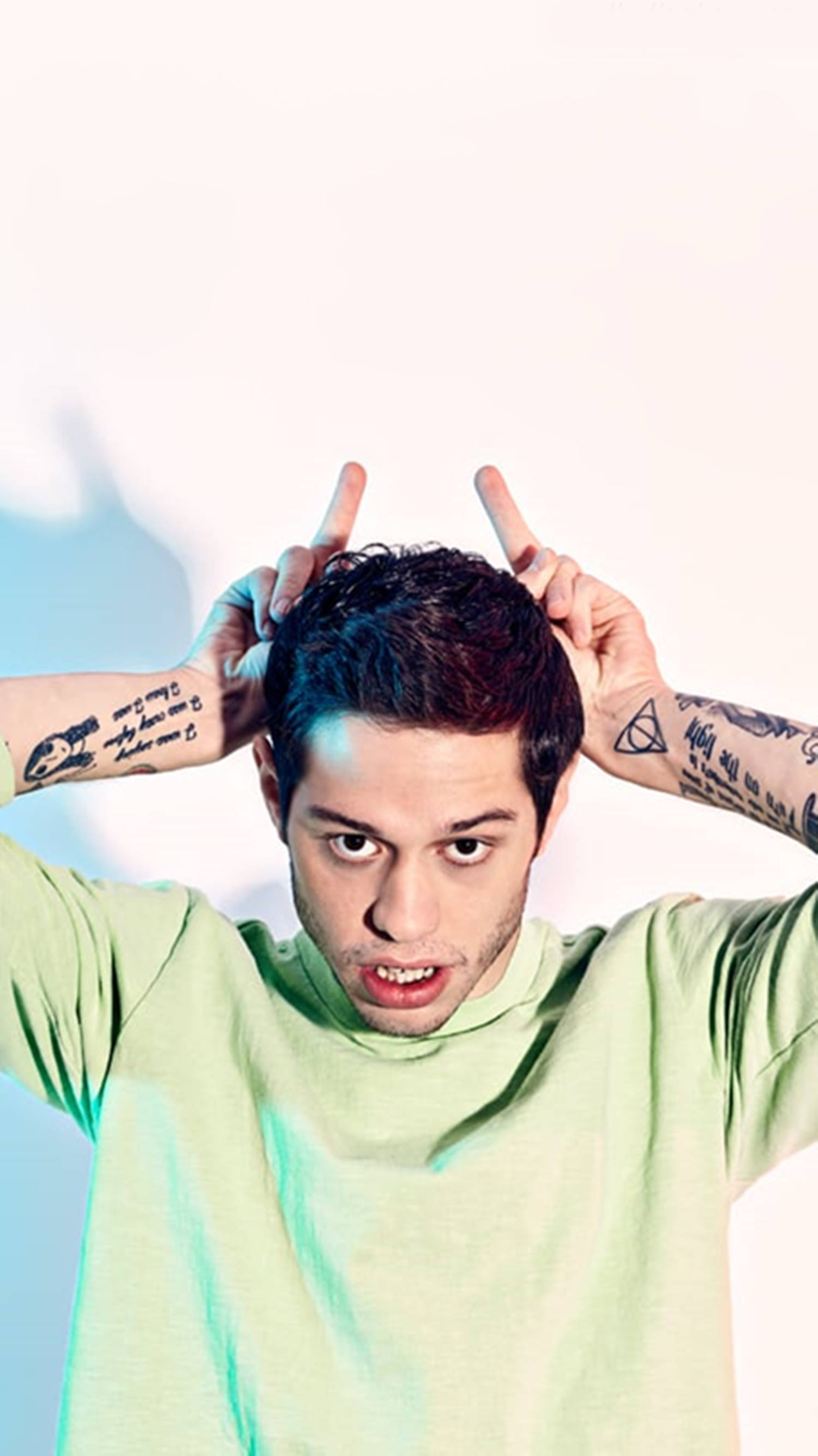 Pete Davidson 在 Complex 雜誌訪問中的肖像照 壁紙 — 一位有紋身的年輕男子在明亮的白色背景前，配合冷色調的藍色燈光，做出魔鬼角手勢。