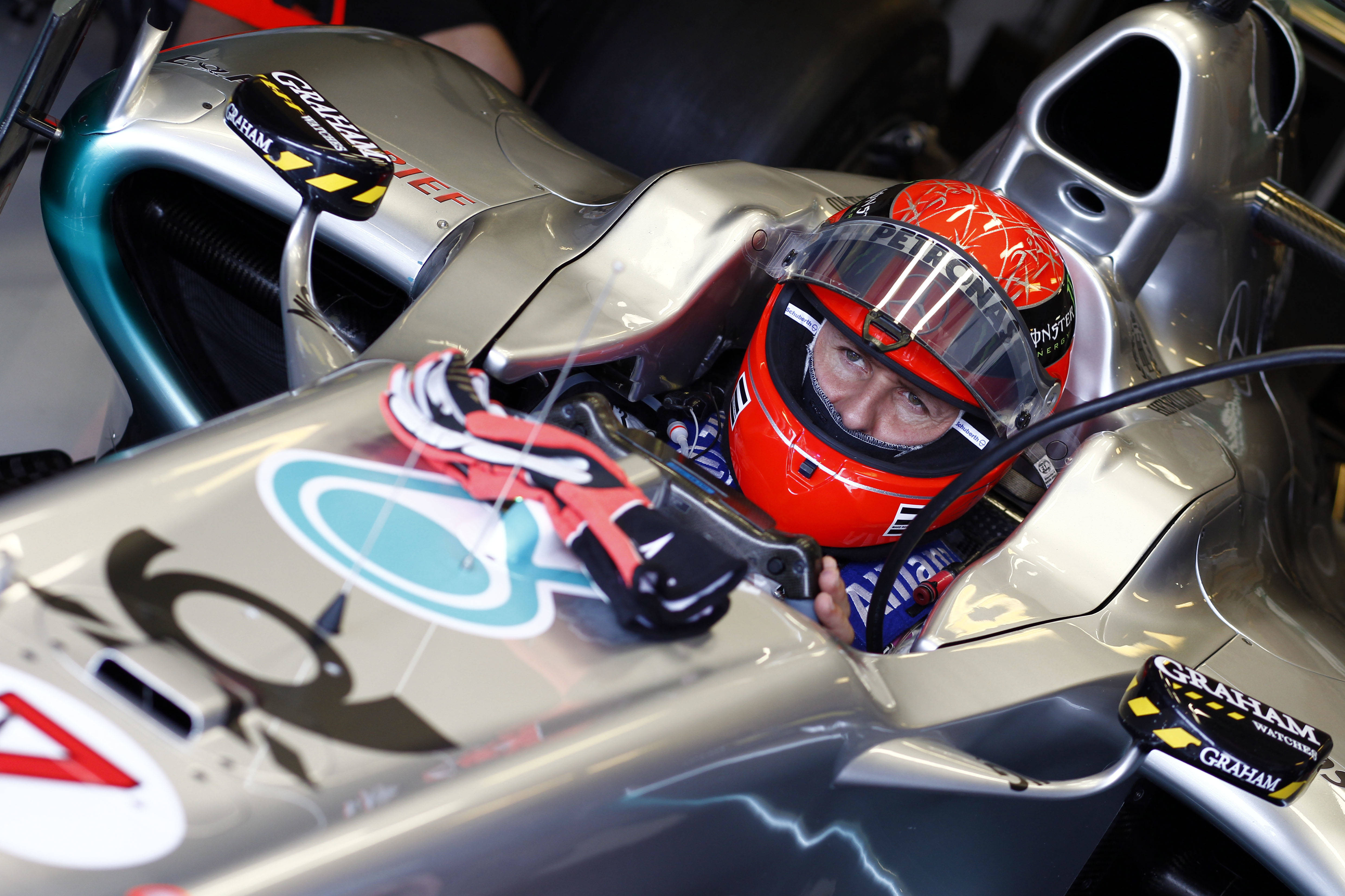 梅賽德斯Petronas舒馬赫賽車壁紙 壁紙 — 一名戴著紅色頭盔的F1車手從銀色和藍綠色的賽車內部專注地凝視，車上有贊助商標誌。