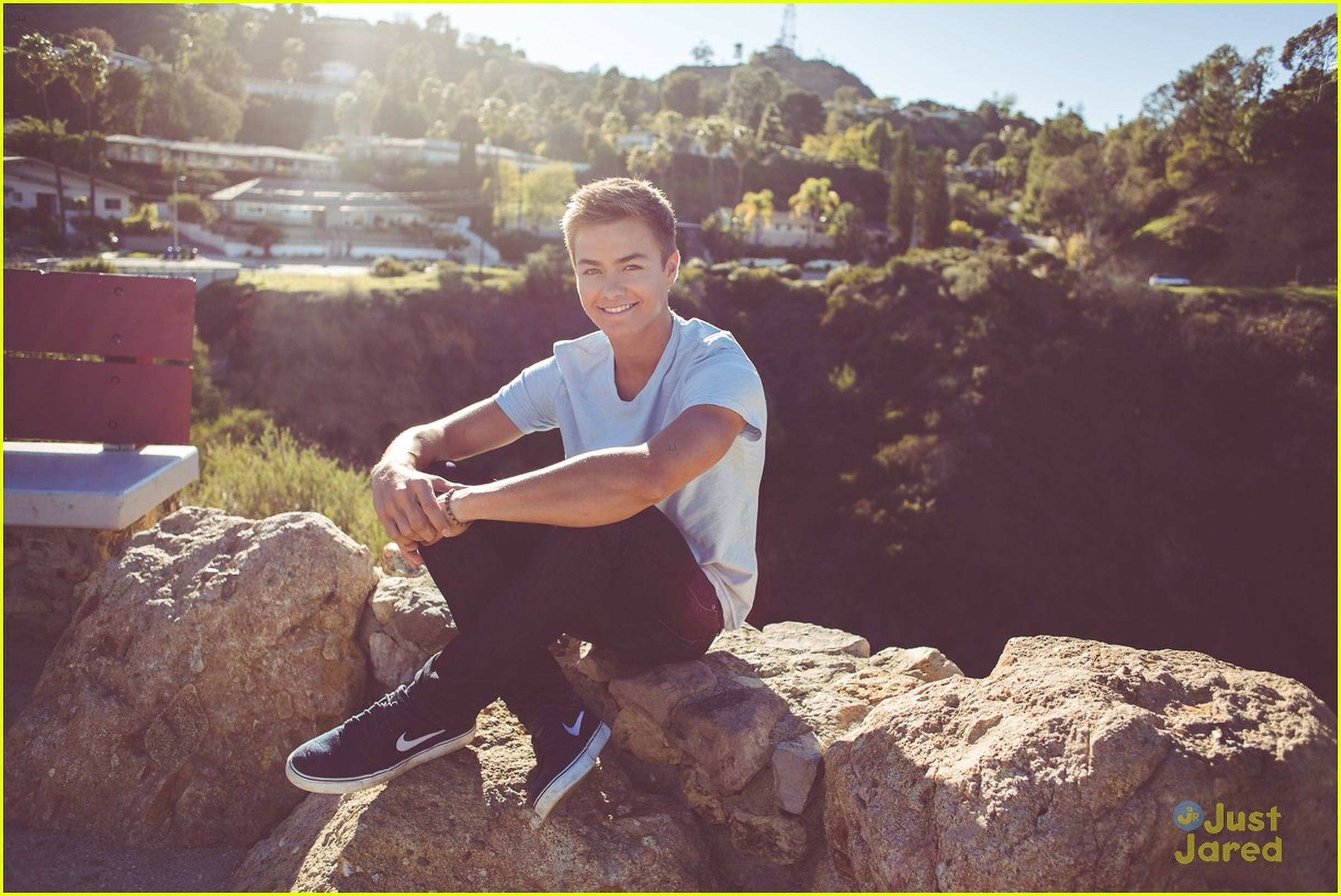 Peyton Meyer Just Jared 專訪拍攝 壁紙 — 一位微笑的年輕男子隨意地坐在陽光普照的岩石上，俯瞰著一個丘陵住宅區，樹木和房屋沐浴在金色的午後陽光中。