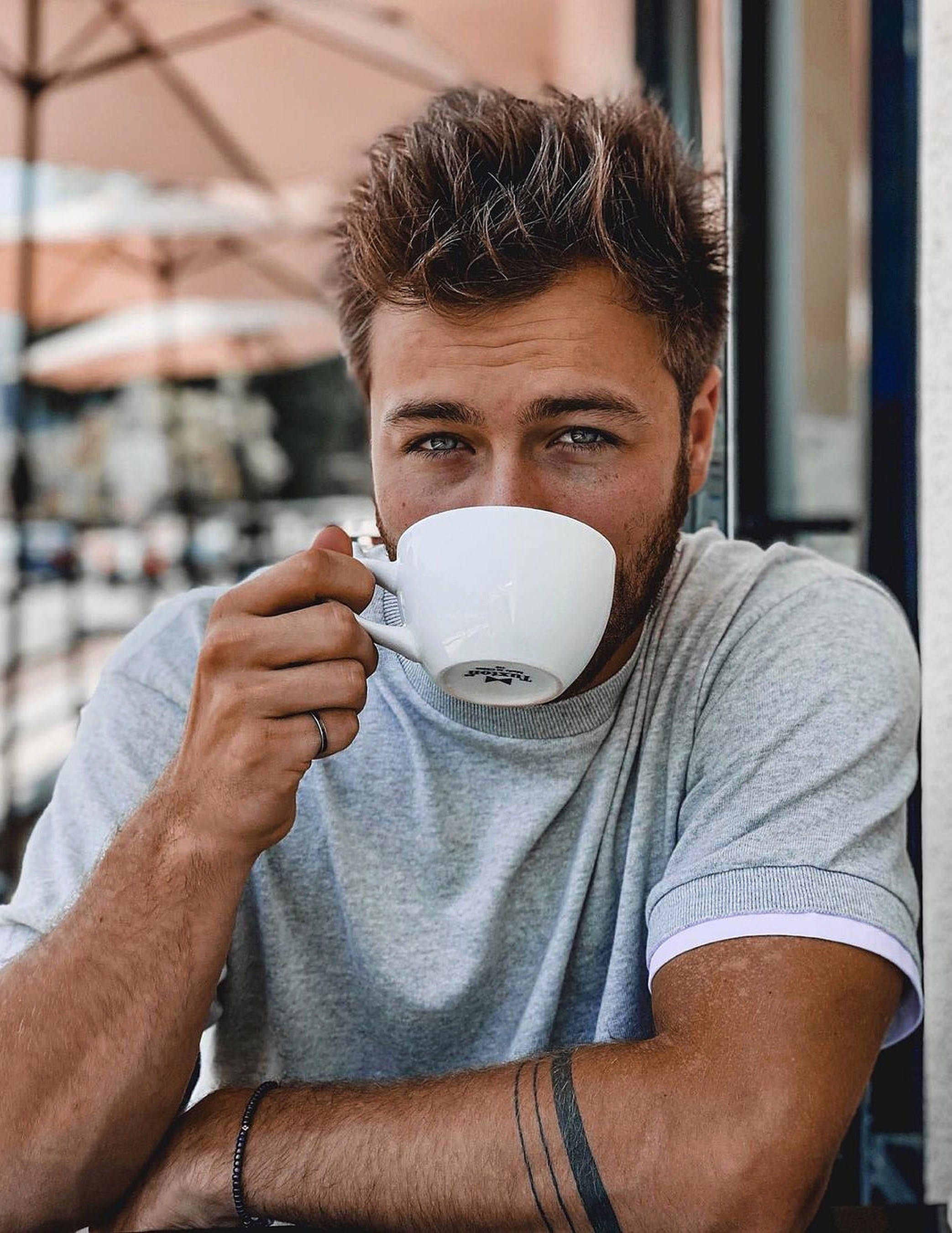 Peyton Meyer 咖啡杯 壁紙 — 一個頭髮凌亂的年輕男子靠著陽光普照的咖啡館窗邊，從白色杯子啜飲咖啡。