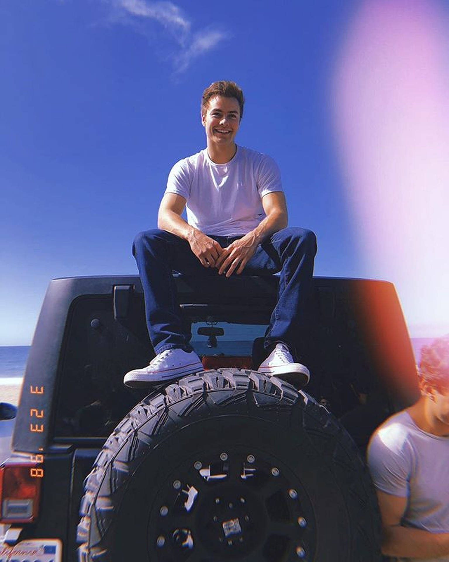 Peyton Meyer 車頂造型 壁紙 — 一個微笑的年輕男子坐在黑色吉普車的備用輪胎上，背景是明亮的藍天和海洋。