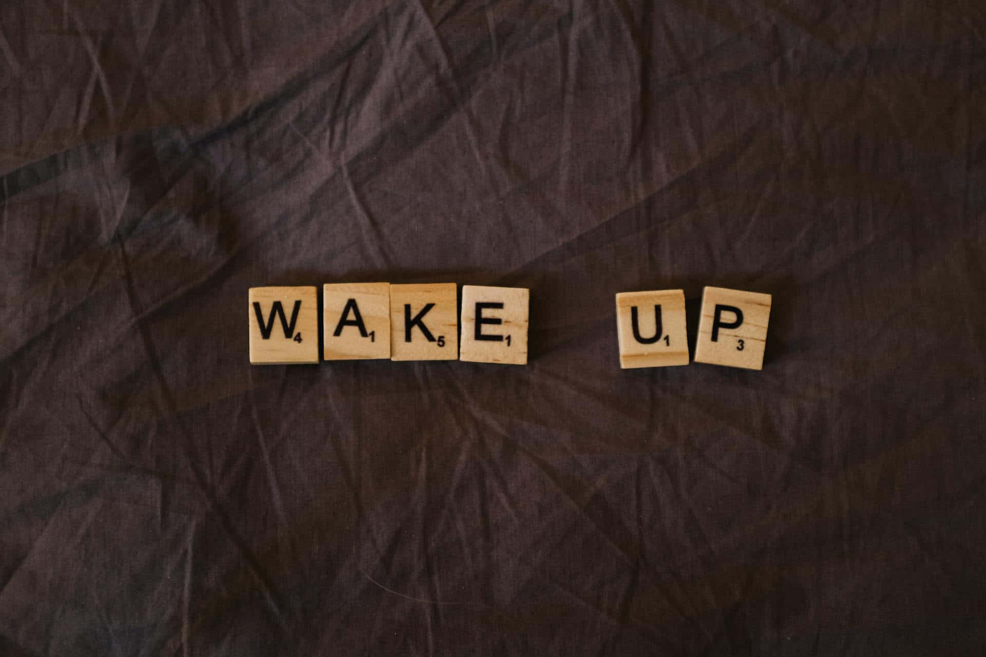  壁紙 — 拼出 WAKE UP 的木製 Scrabble 字母牌放在深色皺褶布料表面上。