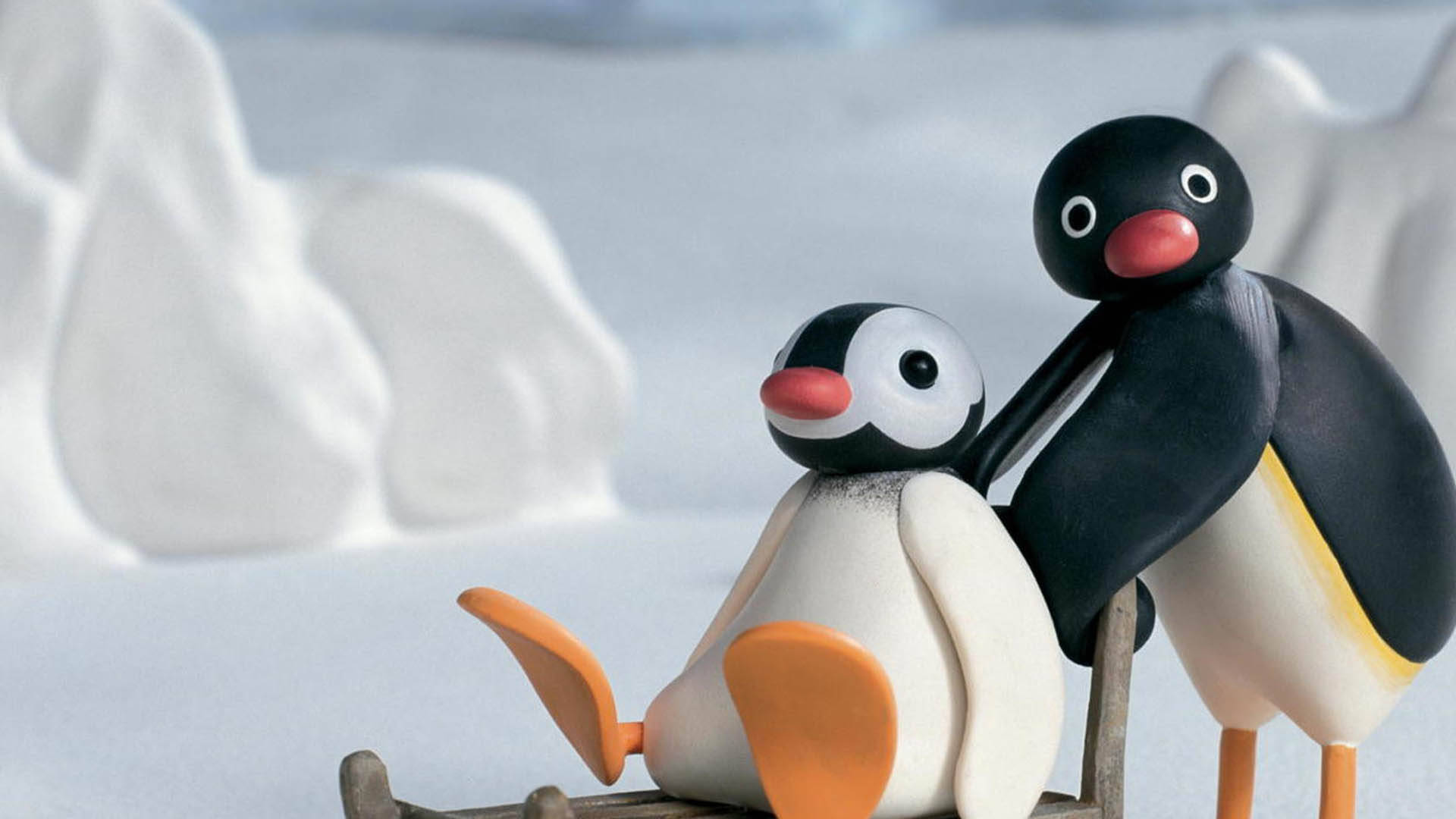 Pingu 與 Pinga 在雪地玩耍 壁紙 — 兩隻黏土動畫企鵝乘坐木製雪橇穿過雪地南極景觀，背景有柔和冰層。