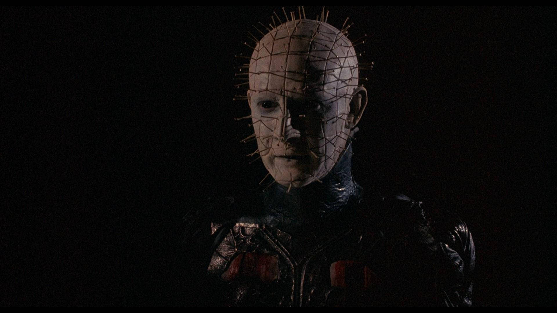 Pinhead 黑暗祭司 壁紙 — 一個帶有尖刺網格圖案頭部和深色皮革服裝的威脅性人物站在陰影黑暗中。