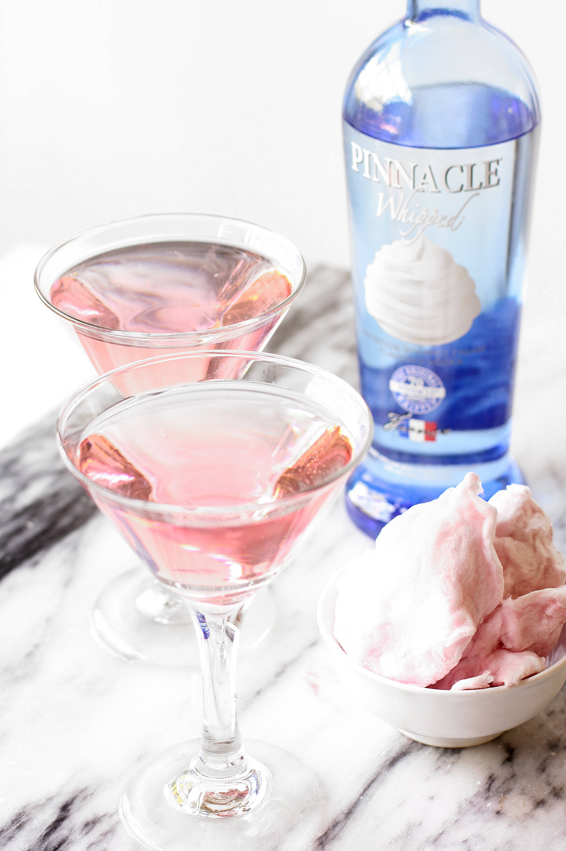 Pinnacle Vodka 馬提尼經典玻璃杯 壁紙 — 兩個粉紅色馬丁尼杯雞尾酒，配以柑橘裝飾，旁邊放著一瓶藍色伏特加和一碗粉紅色棉花糖，置於大理石表面上。