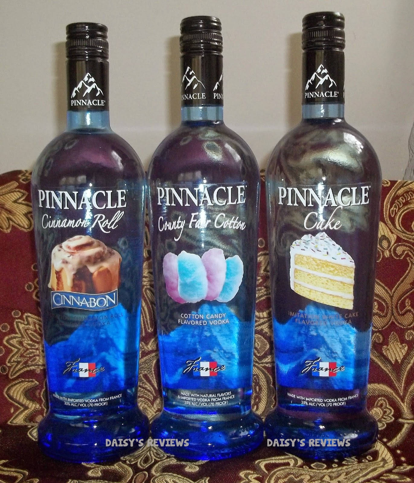 Pinnacle Vodka 肉桂捲、鄉村棉花糖與蛋糕風味 壁紙 — 三瓶藍色Pinnacle調味伏特加並排放置在紅色和金色圖案布料上。