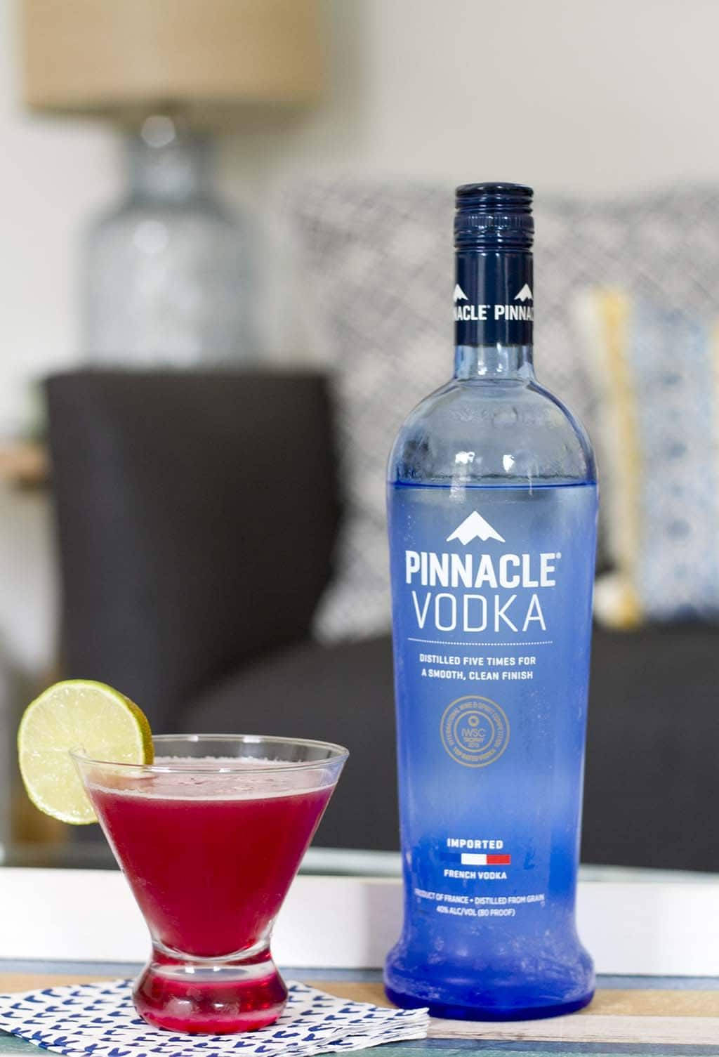 Pinnacle灣畔風味伏特加飲品 壁紙 — 一個充滿活力的藍色伏特加酒瓶旁邊放著一杯深紅色雞尾酒，上面用青檸角裝飾，放在有圖案的杯墊上。