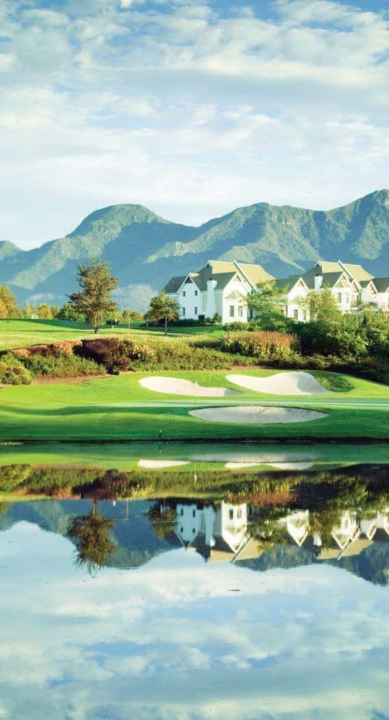 Pixel 3 Fancourt Hotel 高爾夫球場背景 壁紙 — 一個寧靜的高爾夫球場，白色房屋和山脈倒映在平靜的水面上，天空明亮。