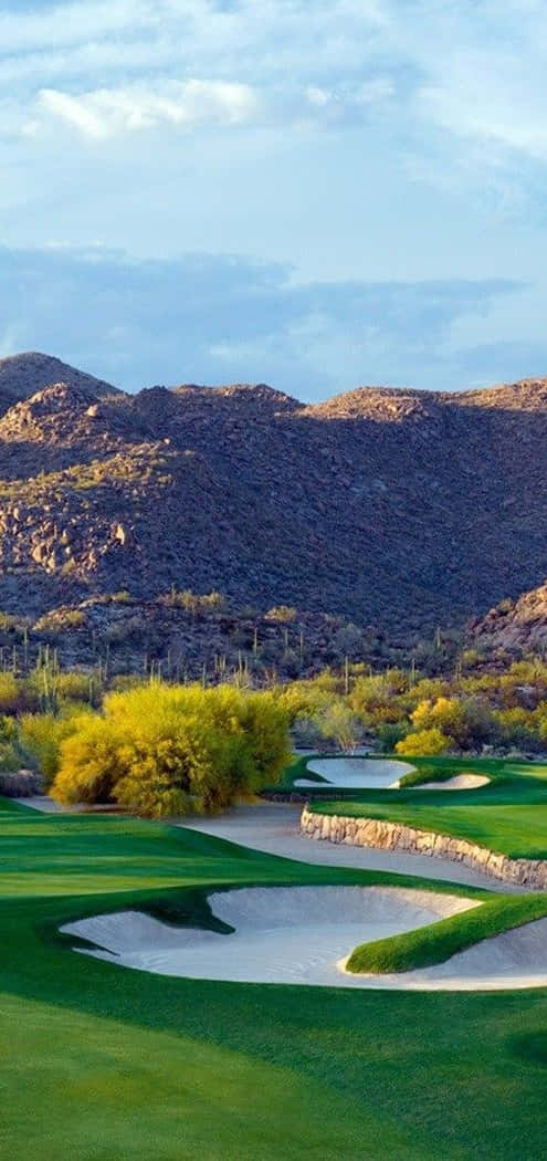 Pixel 3 Renegade 高爾夫球場背景 壁紙 — 一個充滿活力的沙漠高爾夫球場，擁有鬱鬱蔥蔥的綠色球道和沙坑，坐落在崎嶇的山脈之間，天空部分多雲。
