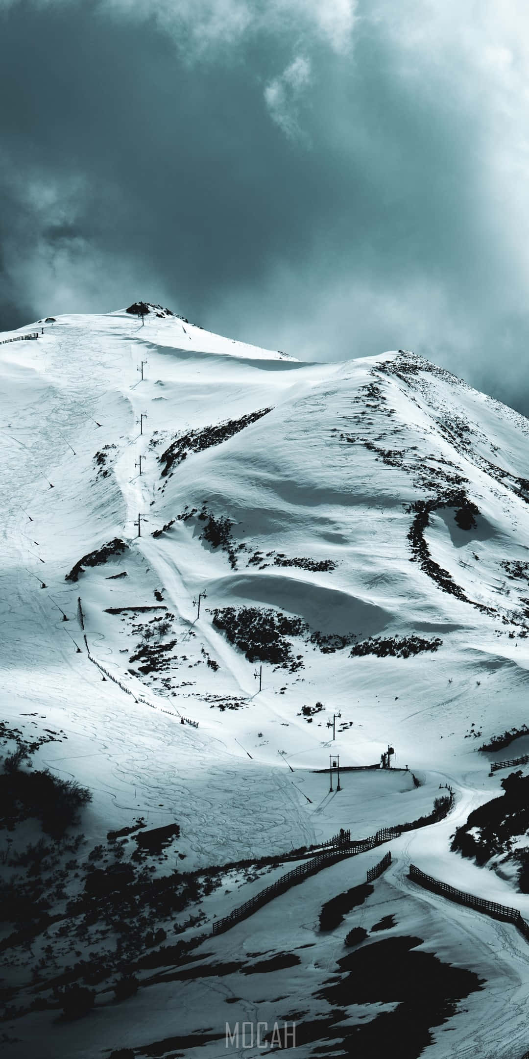 Pixel 3 冬季背景：覆雪山丘 壁紙 — 白雪覆蓋的山峰，天空多雲而戲劇化，設有滑雪纜車和蜿蜒的雪道。