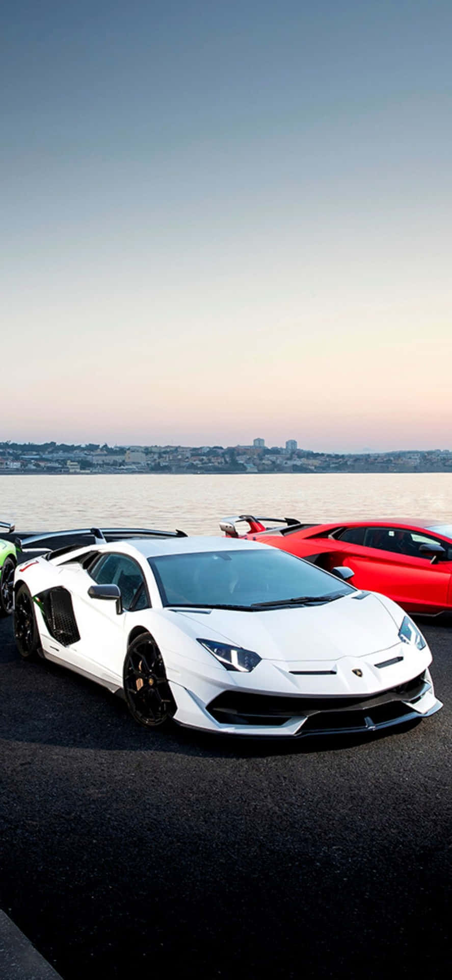 力量與風格：Pixel 3XL Lamborghini 壁紙 — 三輛豪華跑車在日落時停在水邊，背景是城市天際線。