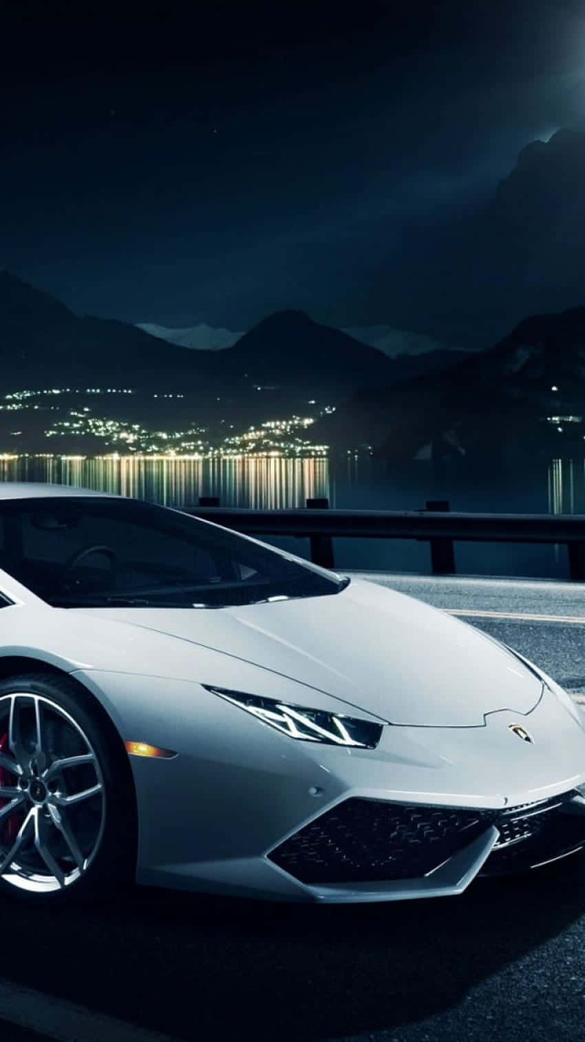 以 Pixel 3XL Lamborghini 風格展現時尚品味 壁紙 — 一輛時尚的銀色跑車在夜燈下閃耀，旁邊是反射的湖泊，遠處的山城在背景中閃爍。