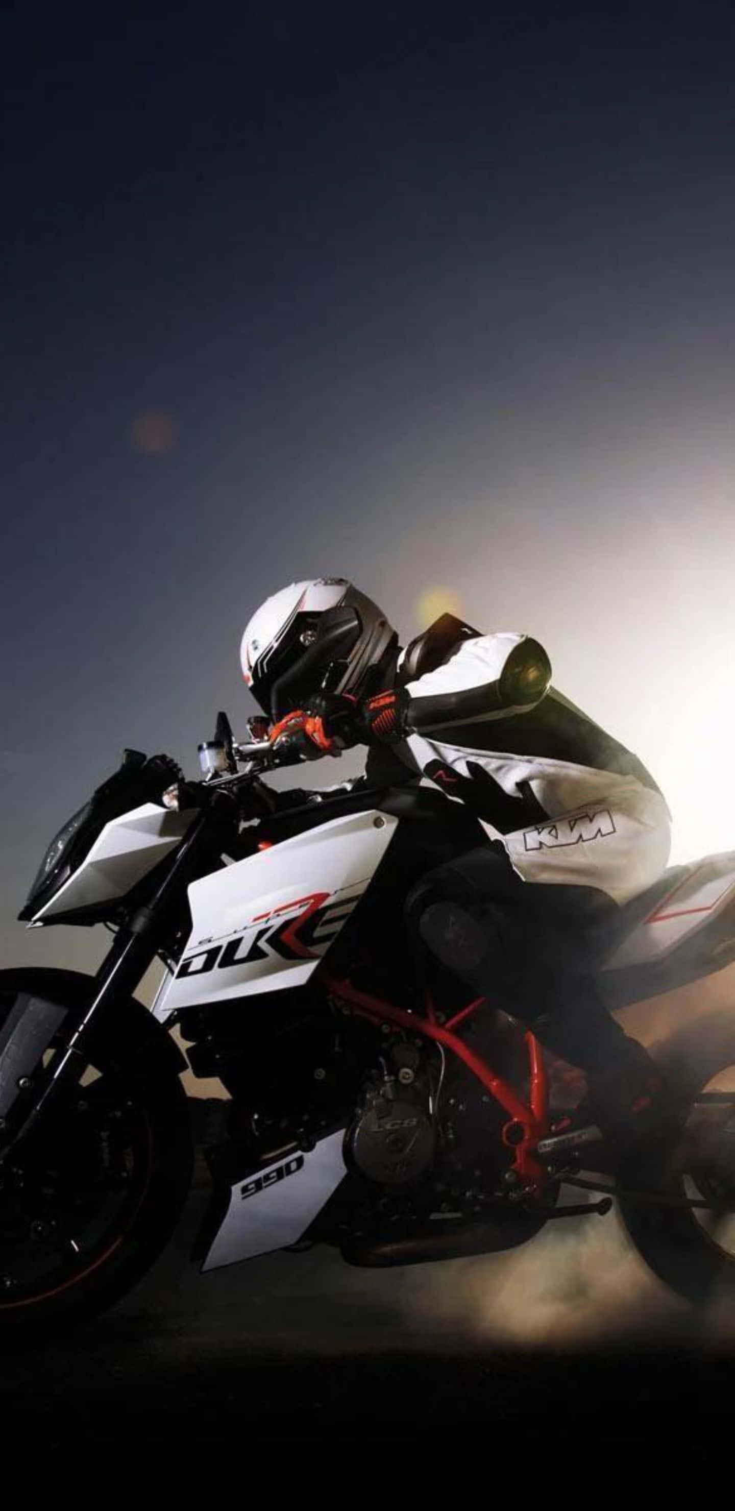 享受順暢騎乘，Pixel 3XL 自行車！ 壁紙 — 一名騎士在白色和紅色的KTM Duke 990電單車上傾斜入彎，背景是黑暗的天空，帶有戲劇性的背光效果。
