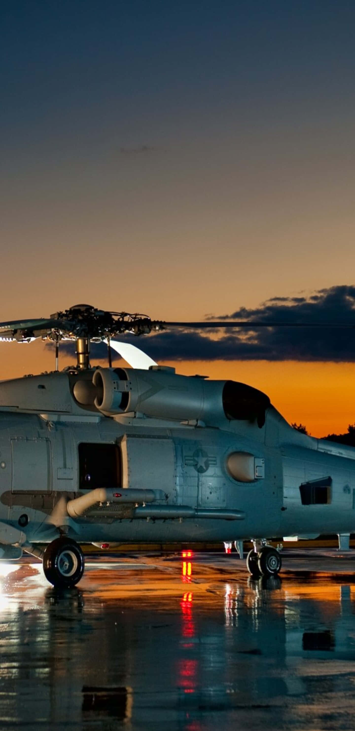 Pixel 3XL 直升機背景 停泊的Sikorsky SH-60 Seahawk 壁紙 — 一架軍用直升機停在濕滑的停機坪上，夕陽西下，鮮豔的橙色和藍色天空倒映在水坑中。