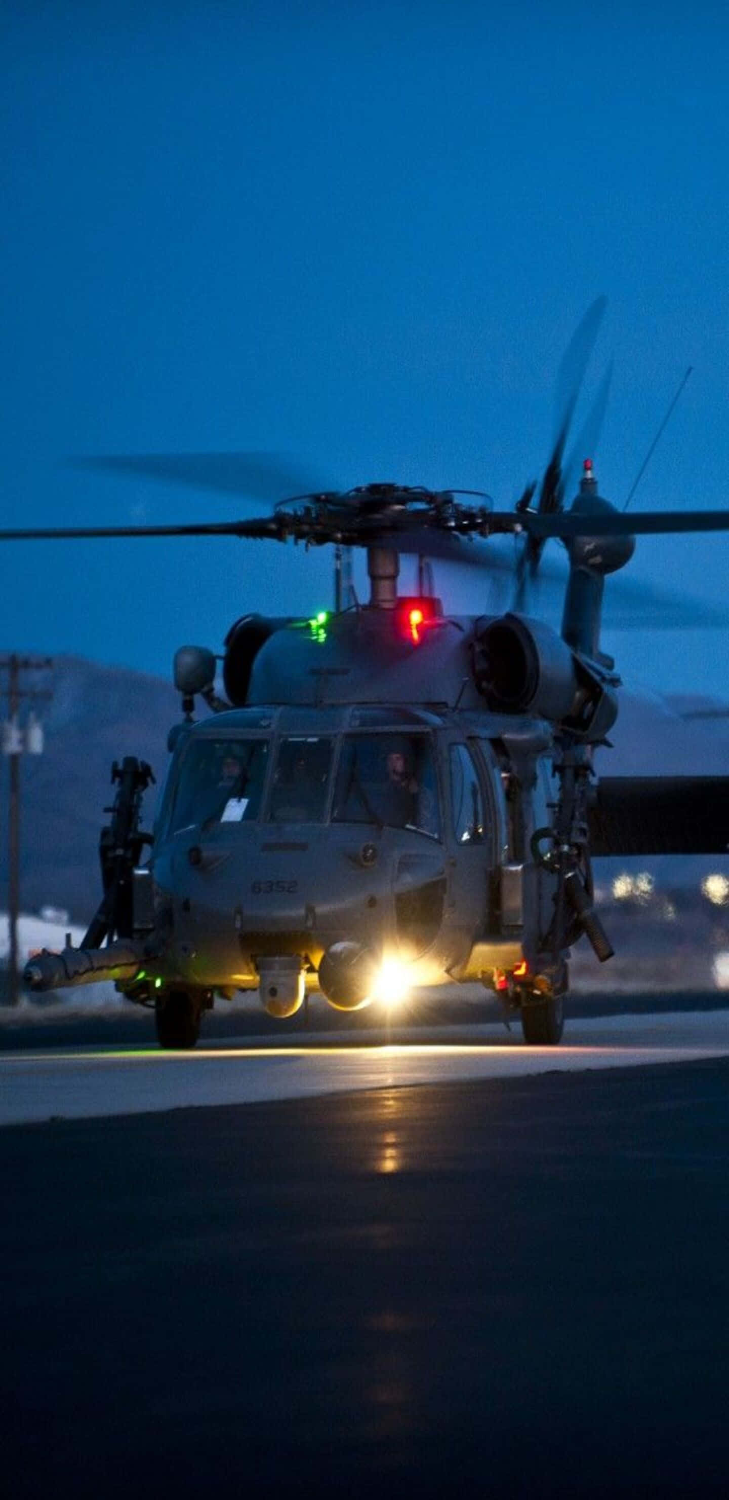 Pixel 3XL 背景：夜間停泊的 Sikorsky UH-60 Black Hawk 直昇機 壁紙 — 一架帶有發光燈光的軍用直升機在深藍色暮色天空下的跑道上滑行。