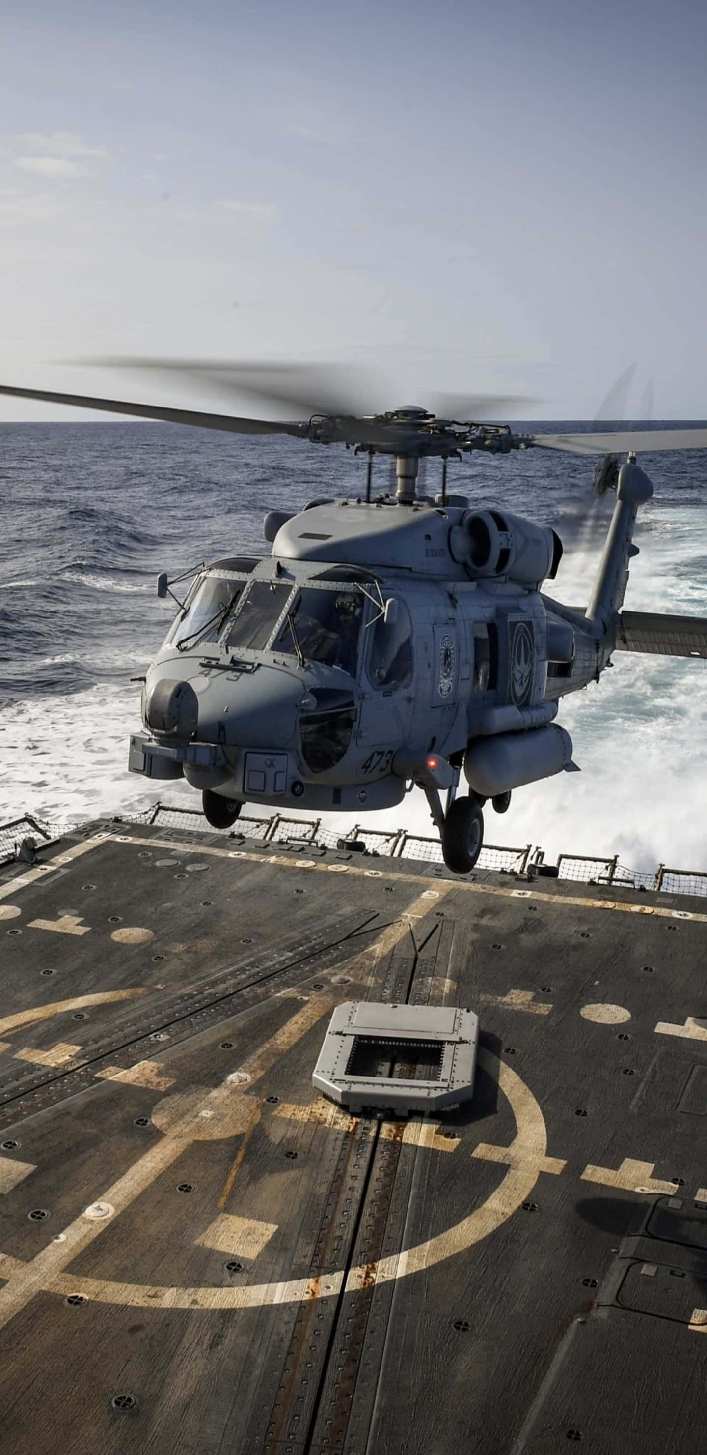 Pixel 3XL 背景：西科斯基 SH-60 海鷹直升機起飛 壁紙 — 一架灰色軍用直升機在軍艦甲板上方盤旋，下方海浪在晴朗天空下拍打。