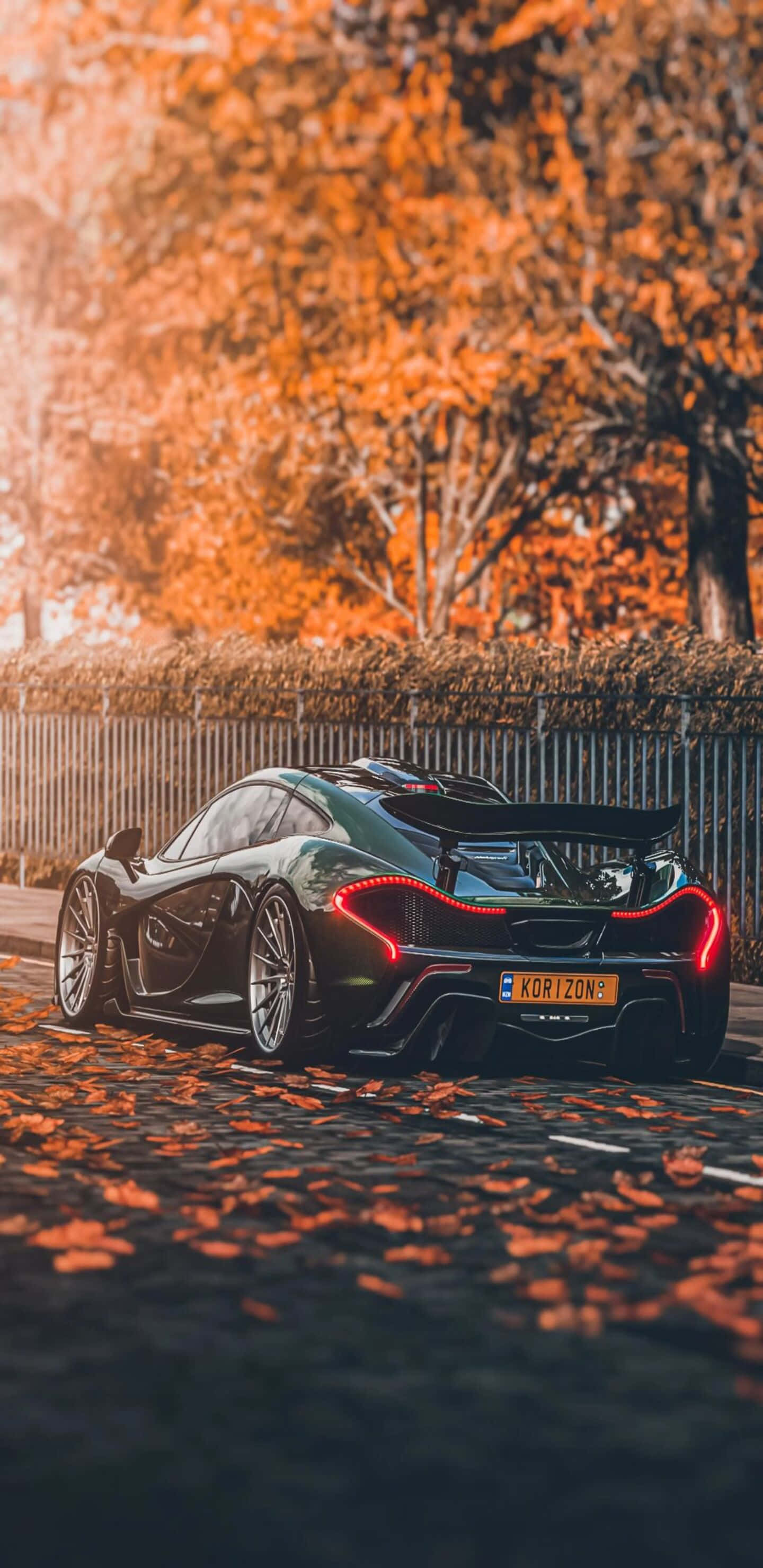 Pixel 3XL 麥拉倫720S Syndicate 背景 壁紙 — 一輛時尚的黑色McLaren跑車，帶有發光的紅色尾燈，停在鋪滿秋葉的鵝卵石街道上，沐浴在溫暖的金色陽光下。